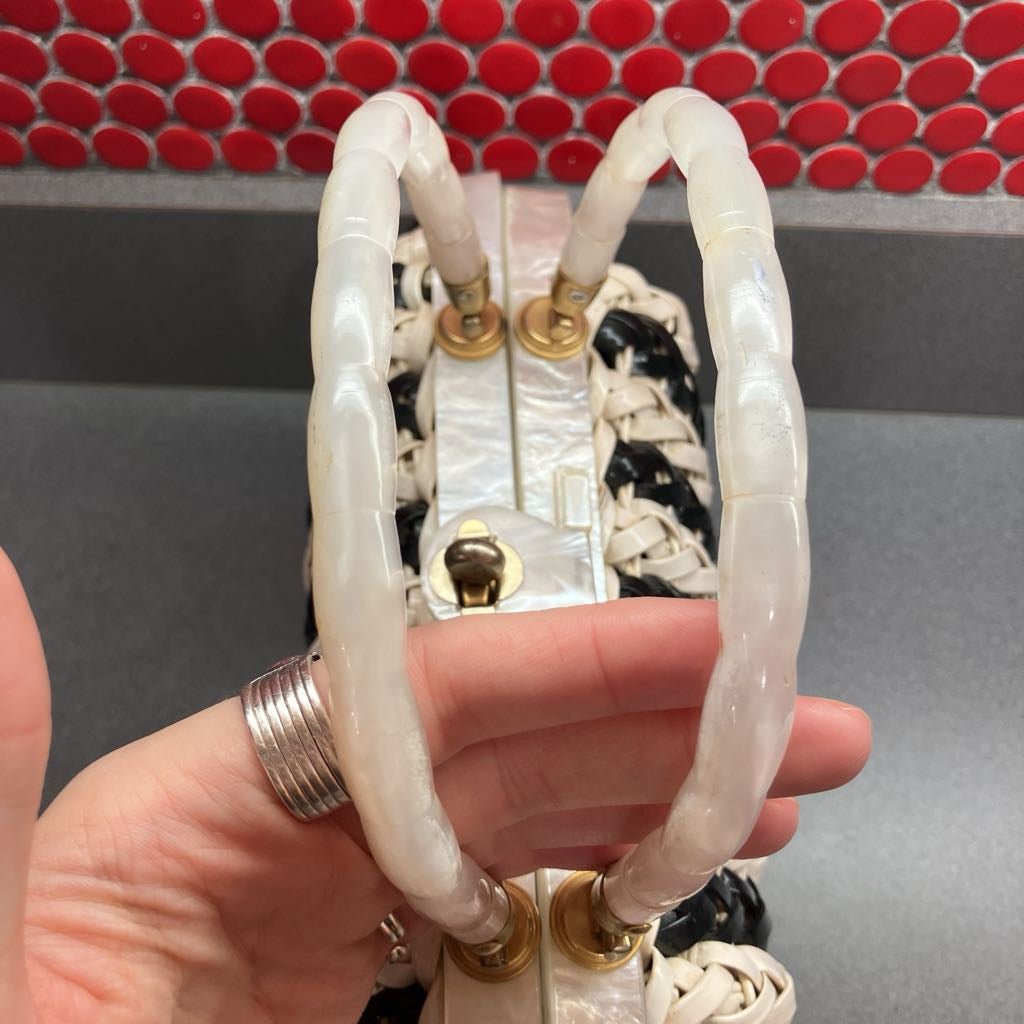 A close-up of a hand holding a woven handbag with chunky, translucent white handles and gold-tone hardware, set against a gray background with a red patterned tile wall.