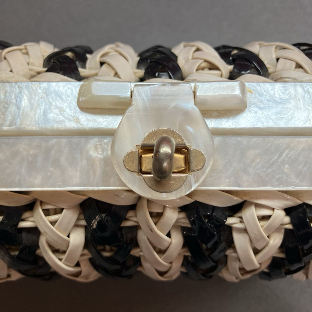 Close-up of a woven clutch bag with cream and black leather straps, a translucent white shell frame, and a gold-tone metal clasp.