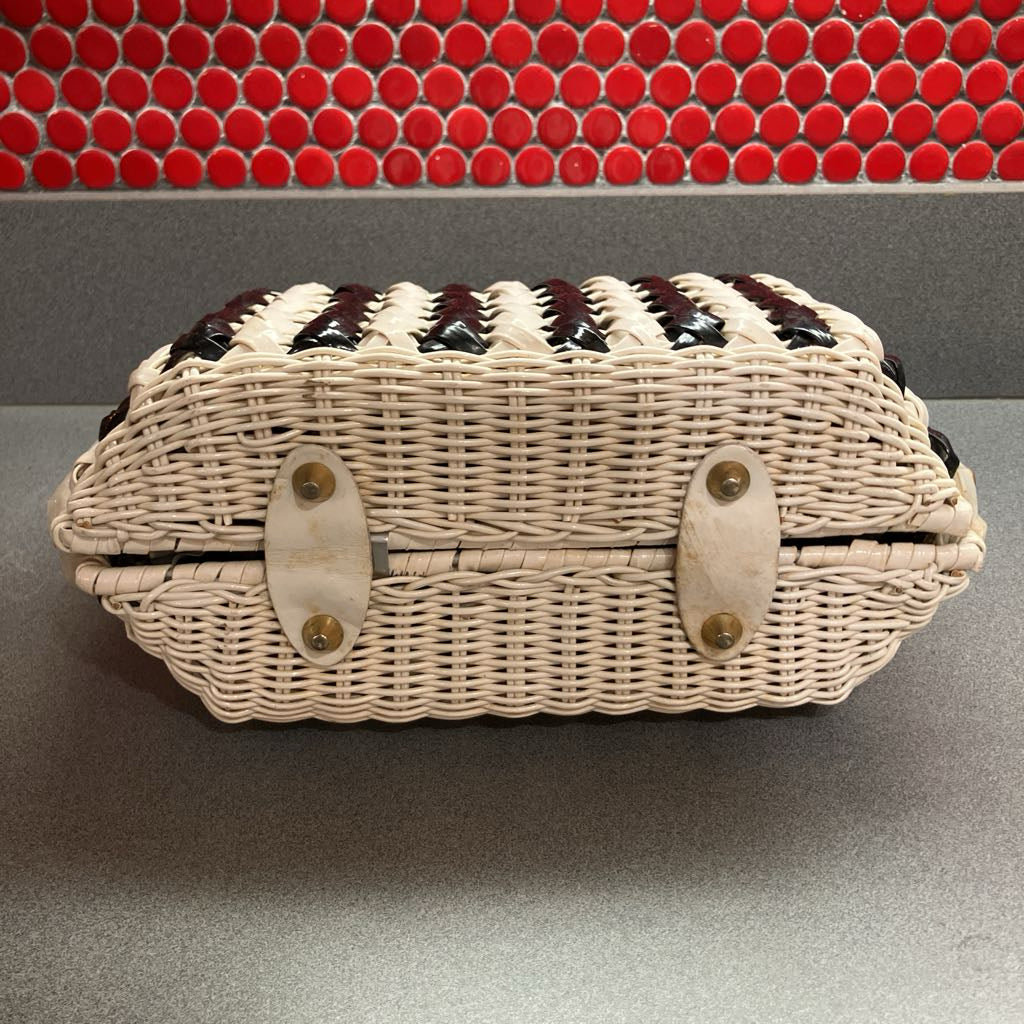 A woven wicker storage basket with a cream-colored body, dark brown chevron accents on the lid, and two cream-colored shell-shaped latches, set against a gray countertop with a red mosaic tile backsplash.