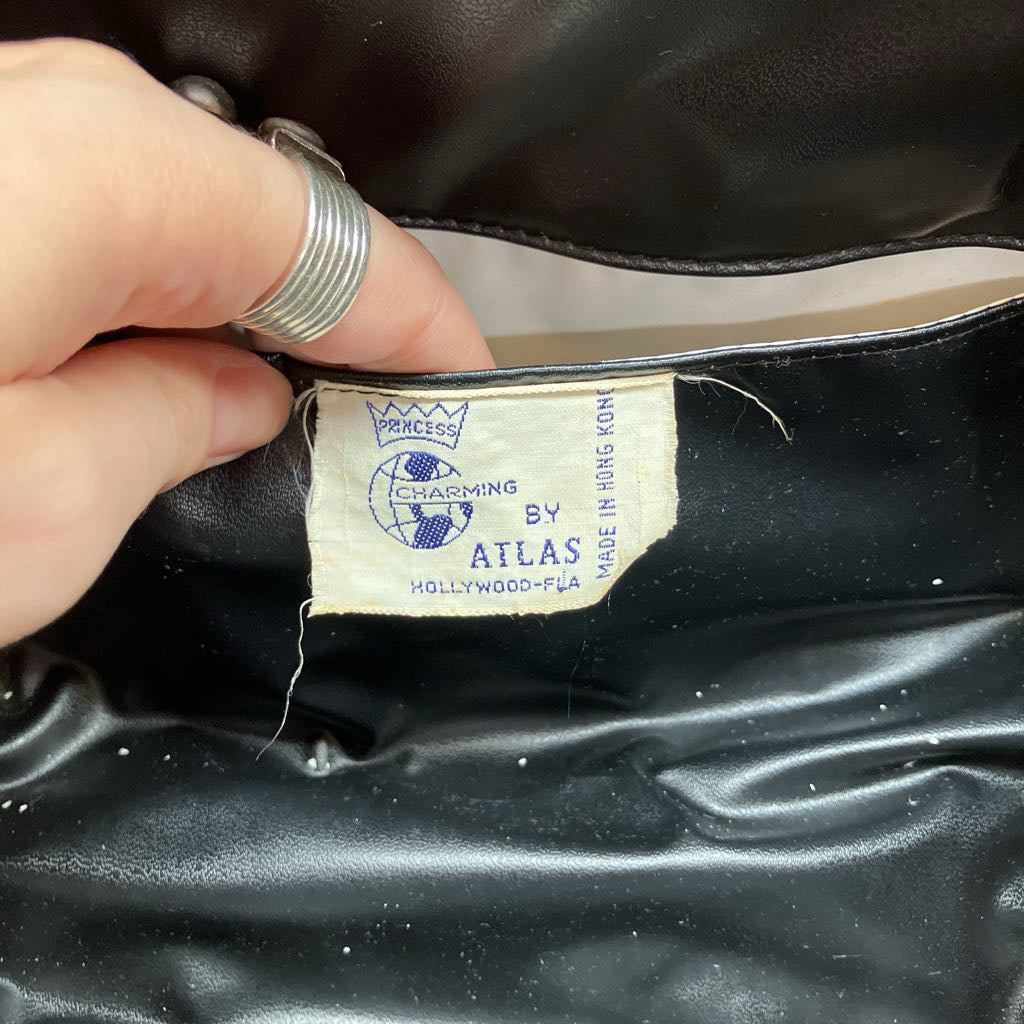 A close-up of a vintage black leather handbag interior showing a beige fabric brand label. A hand with a silver ring is holding the label in place.