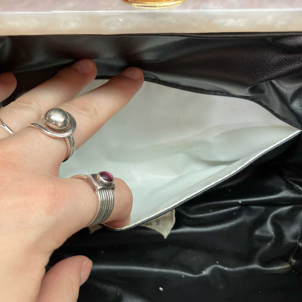 A close-up of a hand wearing silver rings, stretching the opening of a black leather clutch bag to show its white interior lining.