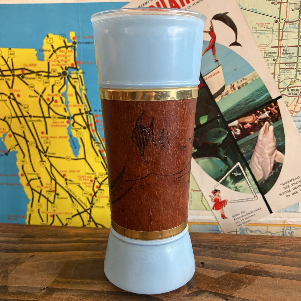 A vintage-style tumbler with a light blue glass top and bottom, a brown faux-wood center wrap, and gold metal bands, standing on a wooden surface with a colorful map collage in the background.