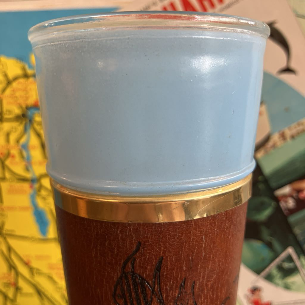 A close-up of a tumbler cup with a light blue frosted top half, a metallic gold band, and a brown textured bottom half with black graphic artwork, set against a colorful background.