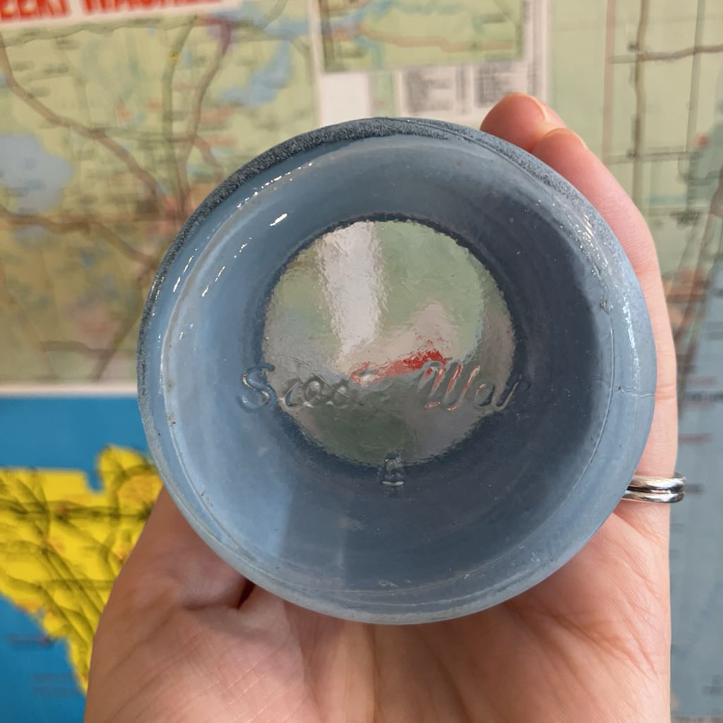 A hand holding the bottom of a light blue ceramic cup or bowl, showing the interior and a stamped brand mark that appears to read 'Siesta Ware.'