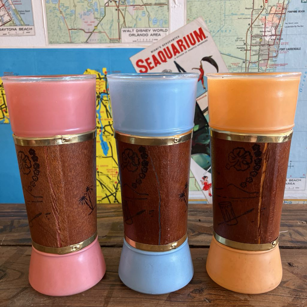 Three vintage-style tumbler cups with wood-grain printed panels and gold bands, shown in pink, blue, and orange colors on a wooden surface with a map and magazine in the background.