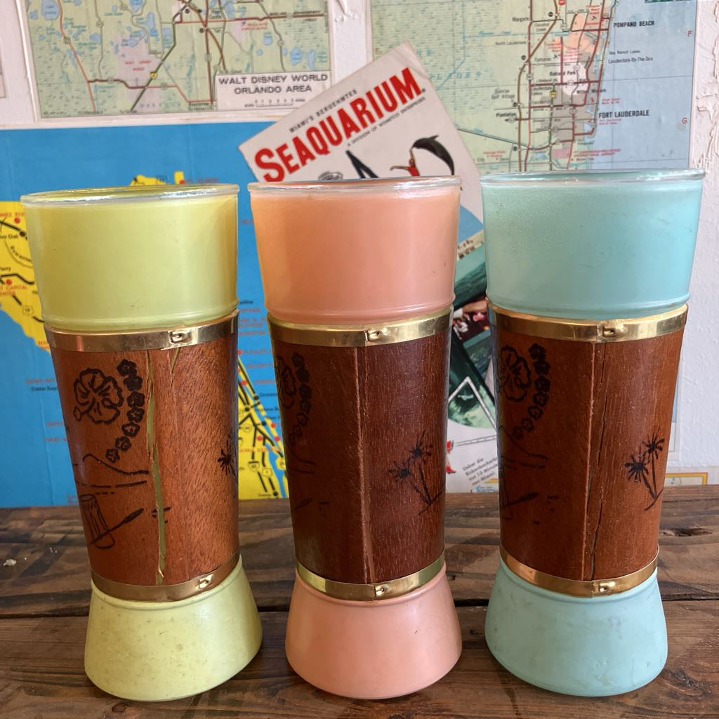 Three vintage-style tumblers with wood-grain panels and gold-tone bands, shown in pale yellow, blush pink, and light blue, standing on a wooden surface with a map and a Seaquarium magazine in the background.
