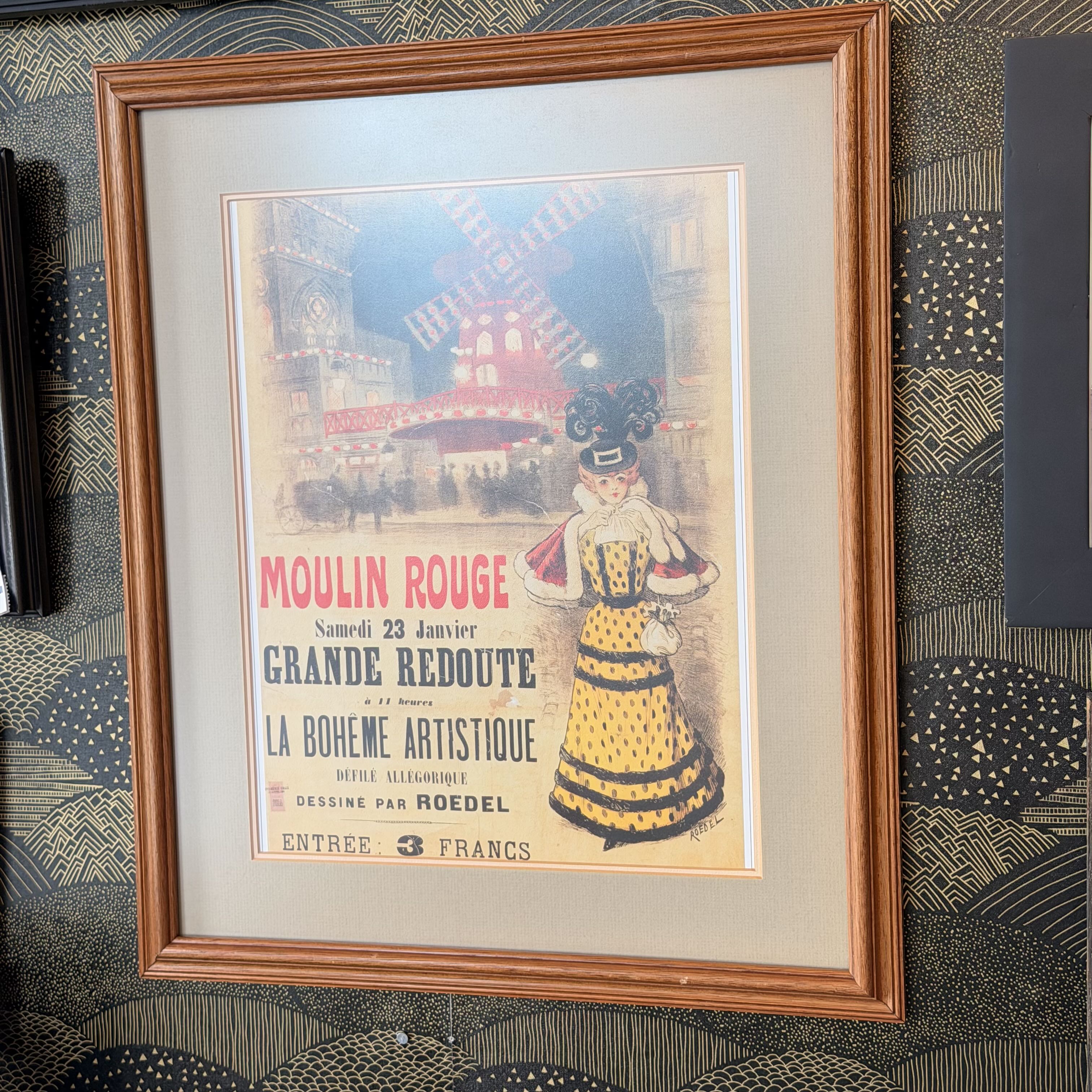 A framed vintage French poster for Moulin Rouge, featuring a woman in a yellow and black dress against a red and blue illuminated stage background, enclosed in a light wood frame and mounted on a patterned wall.