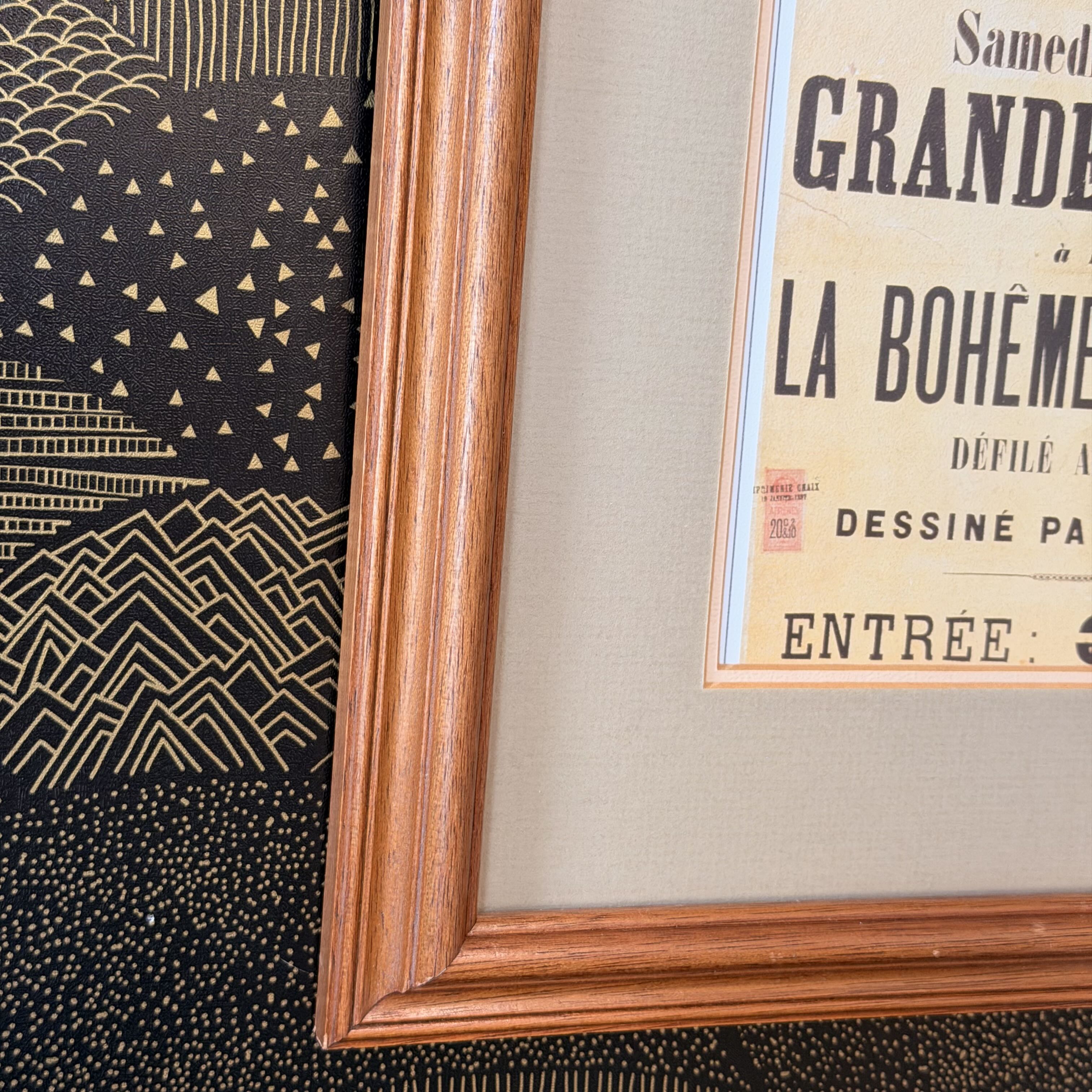 A close-up of a framed art print featuring a vintage French poster with black text on a cream background, set in a warm brown wooden frame against a black and gold patterned backdrop.