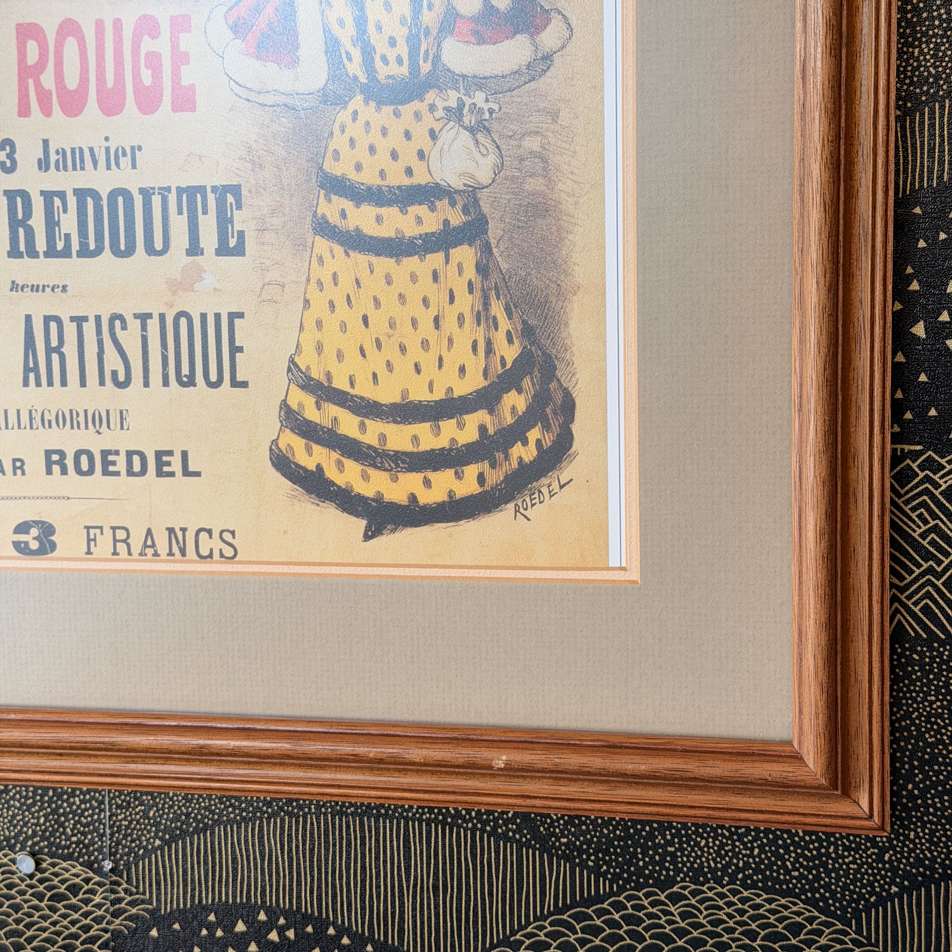 A framed vintage French art print featuring a yellow patterned dress with black stripes and bold red and blue typography, set in a warm brown wood frame with a beige mat.
