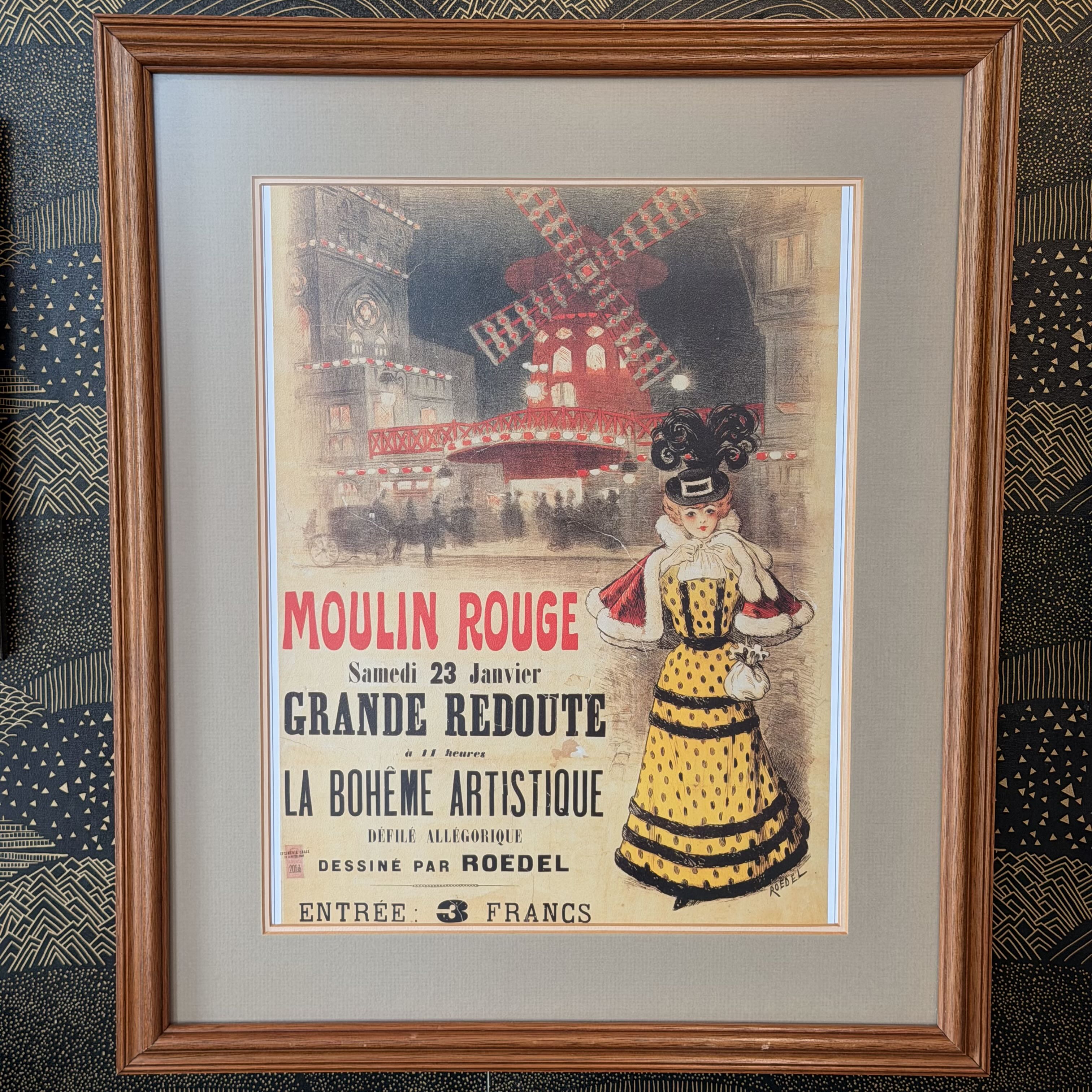 A framed vintage French poster for Moulin Rouge, featuring an illustration of a woman in a yellow dress and a red windmill, set in a brown wooden frame with a gray mat.