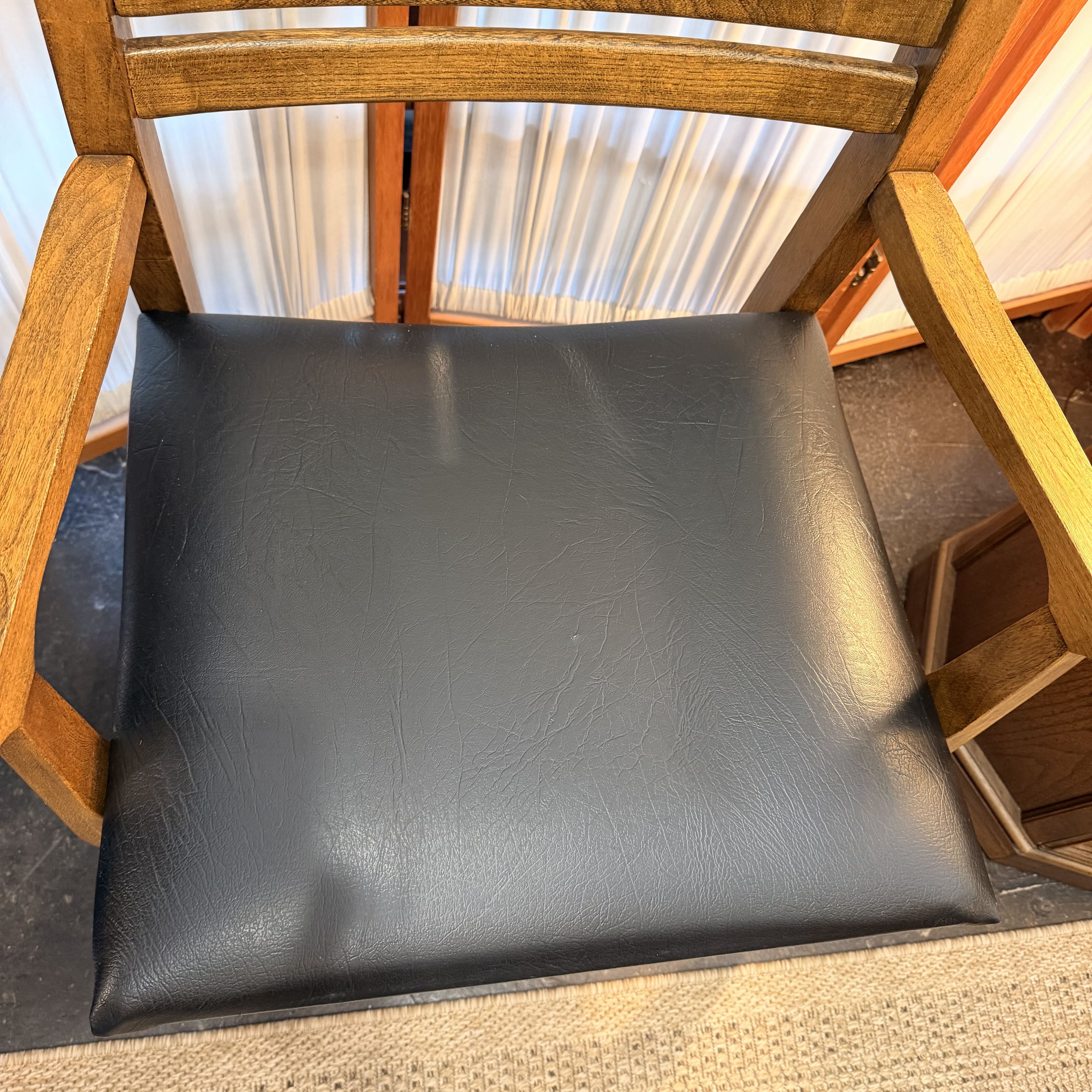 A square black faux-leather or vinyl chair cushion with a subtle pebbled texture, shown installed on a wooden armchair.