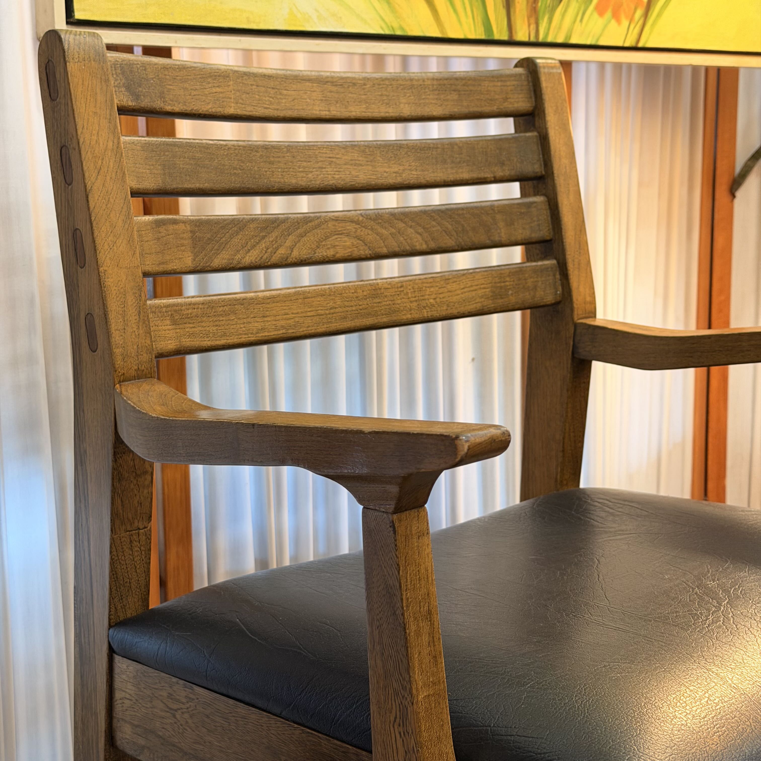 A close-up of a wooden dining chair with a slatted backrest, armrests, and a black upholstered seat cushion, shown indoors against curtains and a colorful painting.