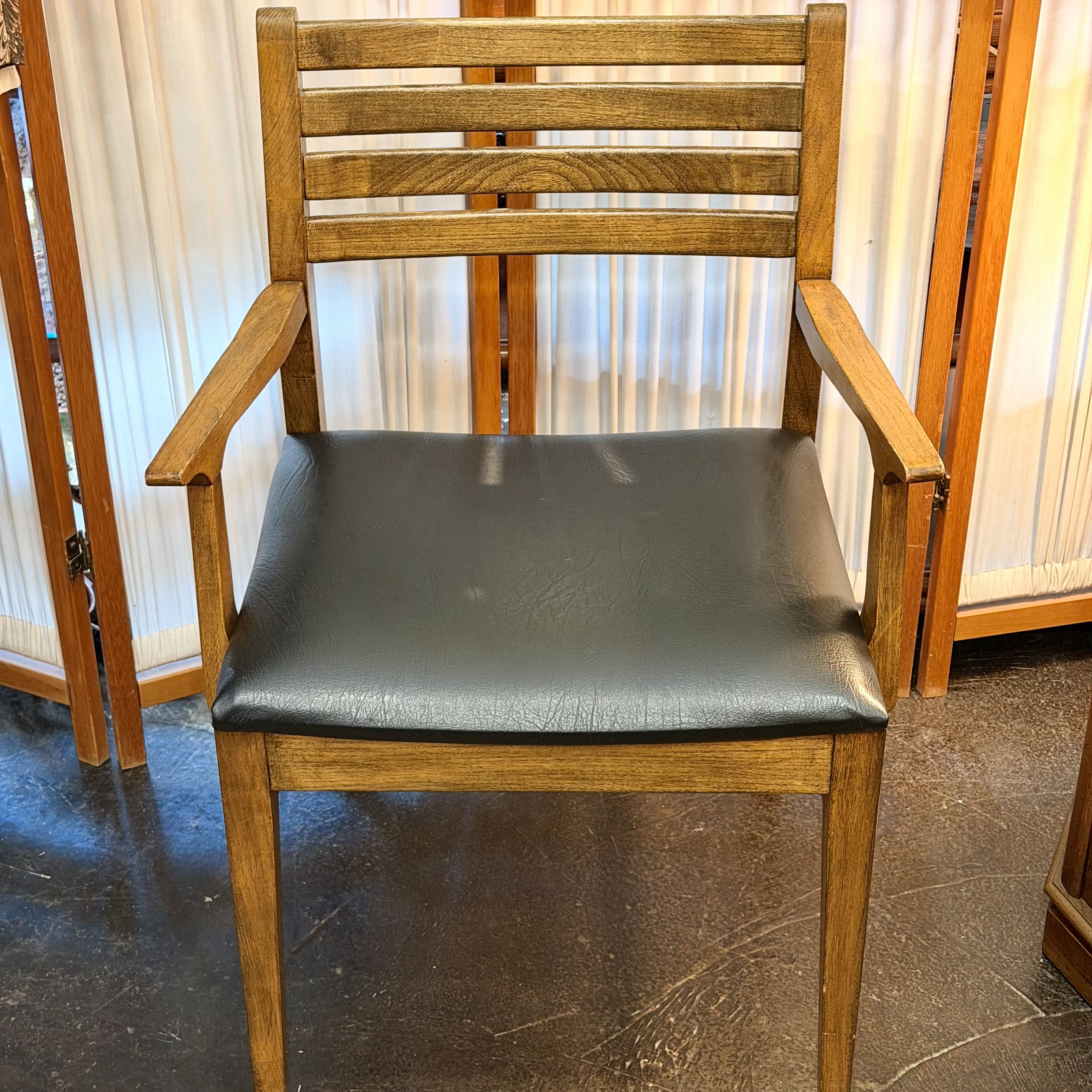 A wooden dining chair with a slatted backrest and armrests, featuring a black faux-leather upholstered seat cushion, shown in an indoor setting.
