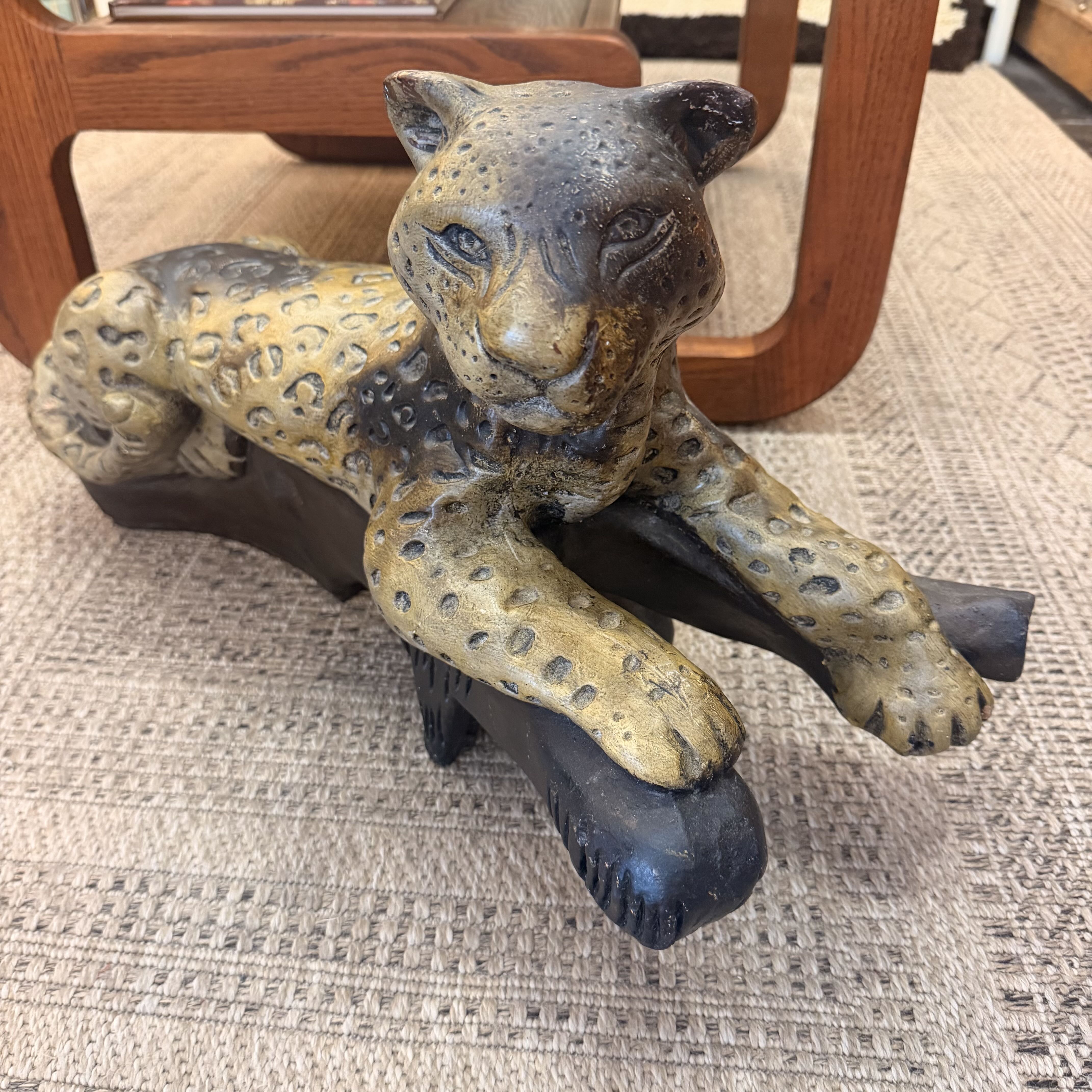 A carved leopard figurine with a spotted tan and dark brown finish, posed lying down on a dark base, photographed indoors on a woven rug with wooden furniture visible in the background.