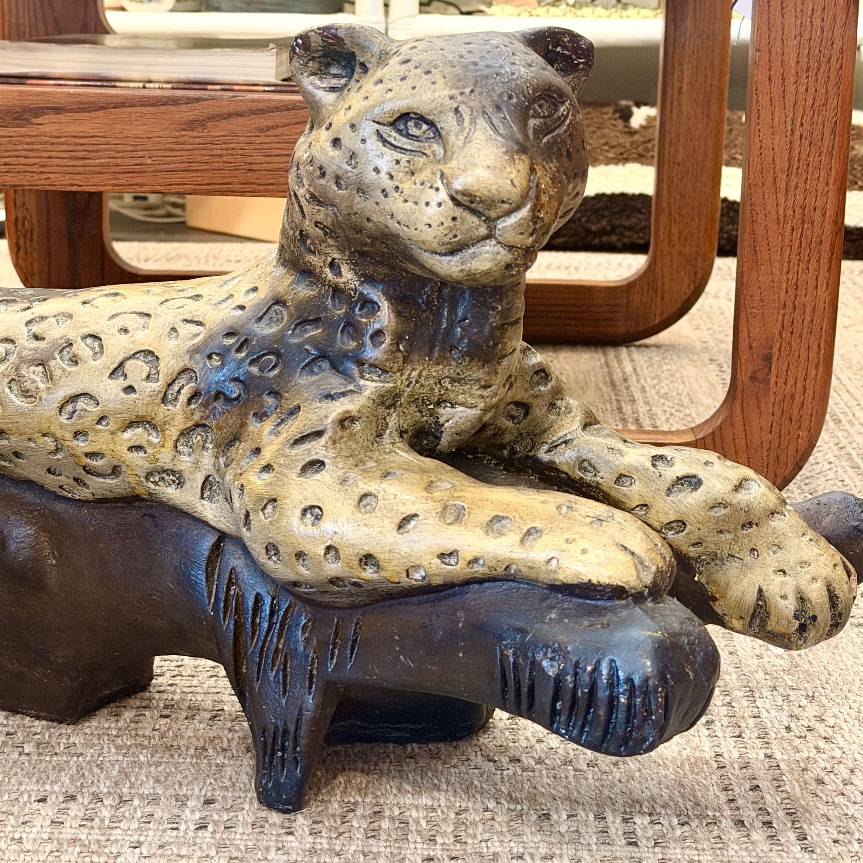 A leopard figurine with a spotted tan and brown coat resting on a dark base, photographed in a real indoor setting with furniture legs visible in the background.