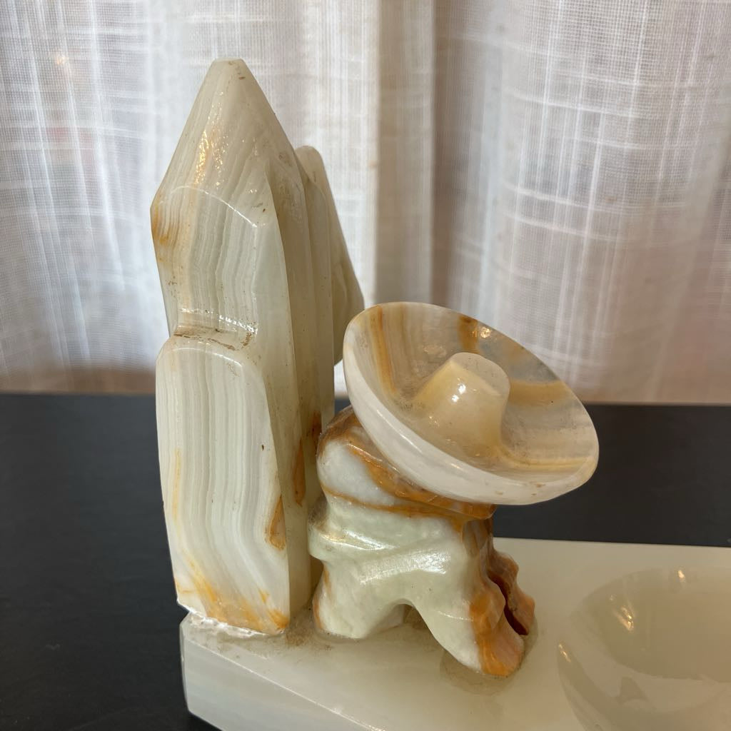 A carved onyx stone sculpture featuring a tall crystal-like tower beside a seated figure with a bowl-shaped head, displayed on a dark tabletop in front of sheer curtains.