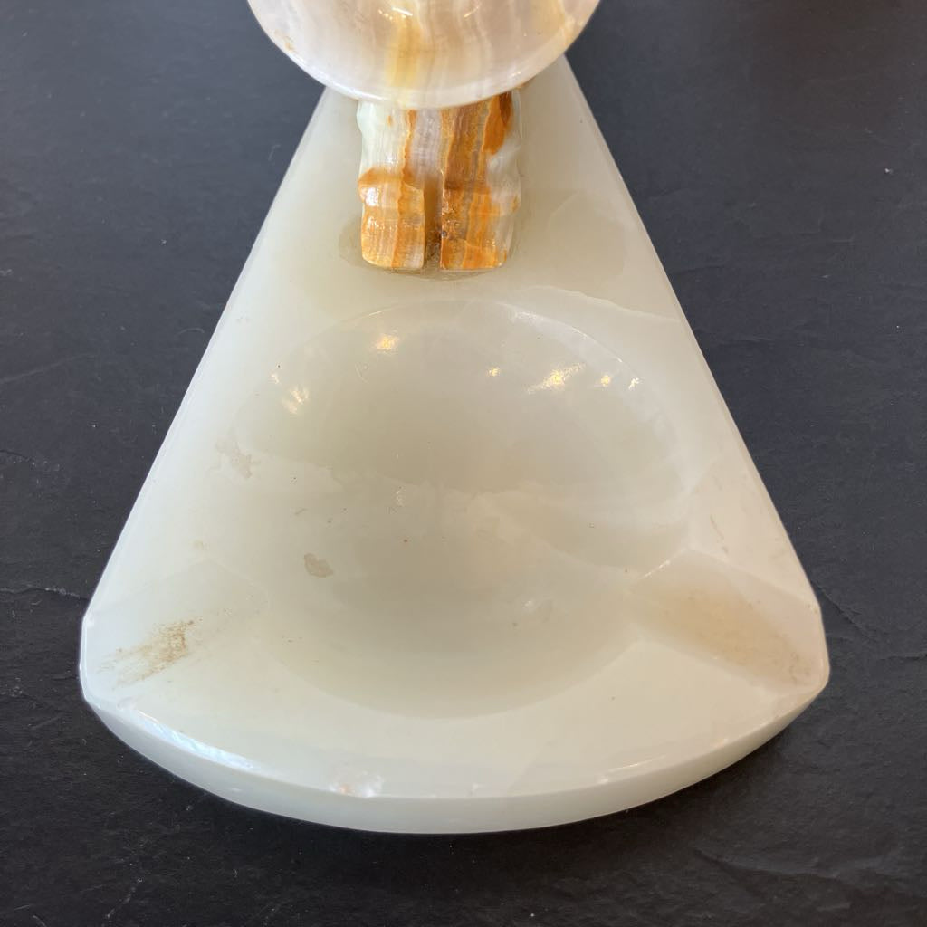A triangular white onyx ashtray with a carved recessed bowl, featuring a matching onyx stand with orange and cream marbling, set against a dark slate background.
