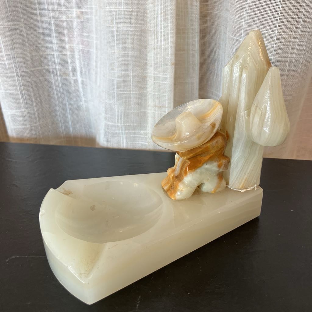 A carved stone decorative object with a shallow oval basin on a rectangular base, alongside sculpted abstract forms in milky white and tan marbled stone, placed on a dark tabletop in front of sheer curtains.