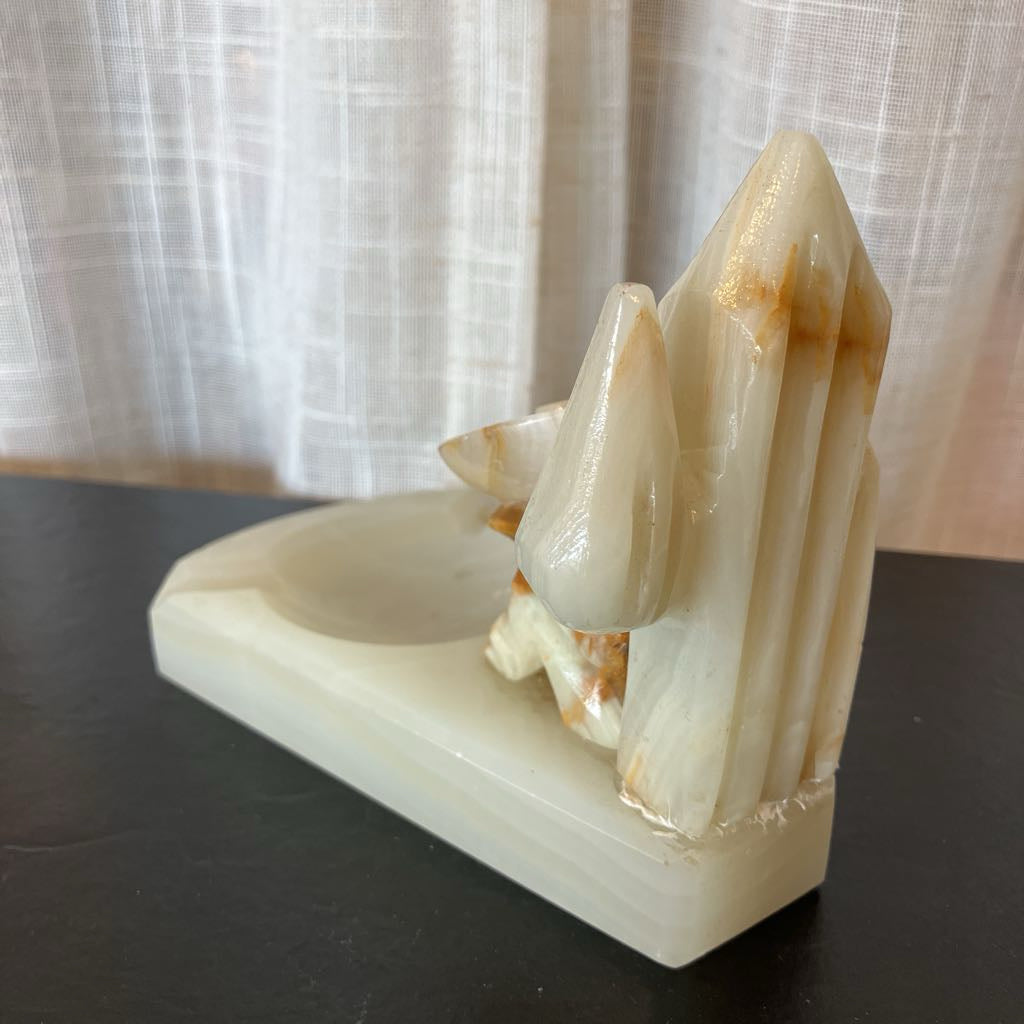 A carved white onyx desk sculpture featuring a stylized animal and tall crystal-like peaks, displayed on a dark tabletop in front of sheer curtains.