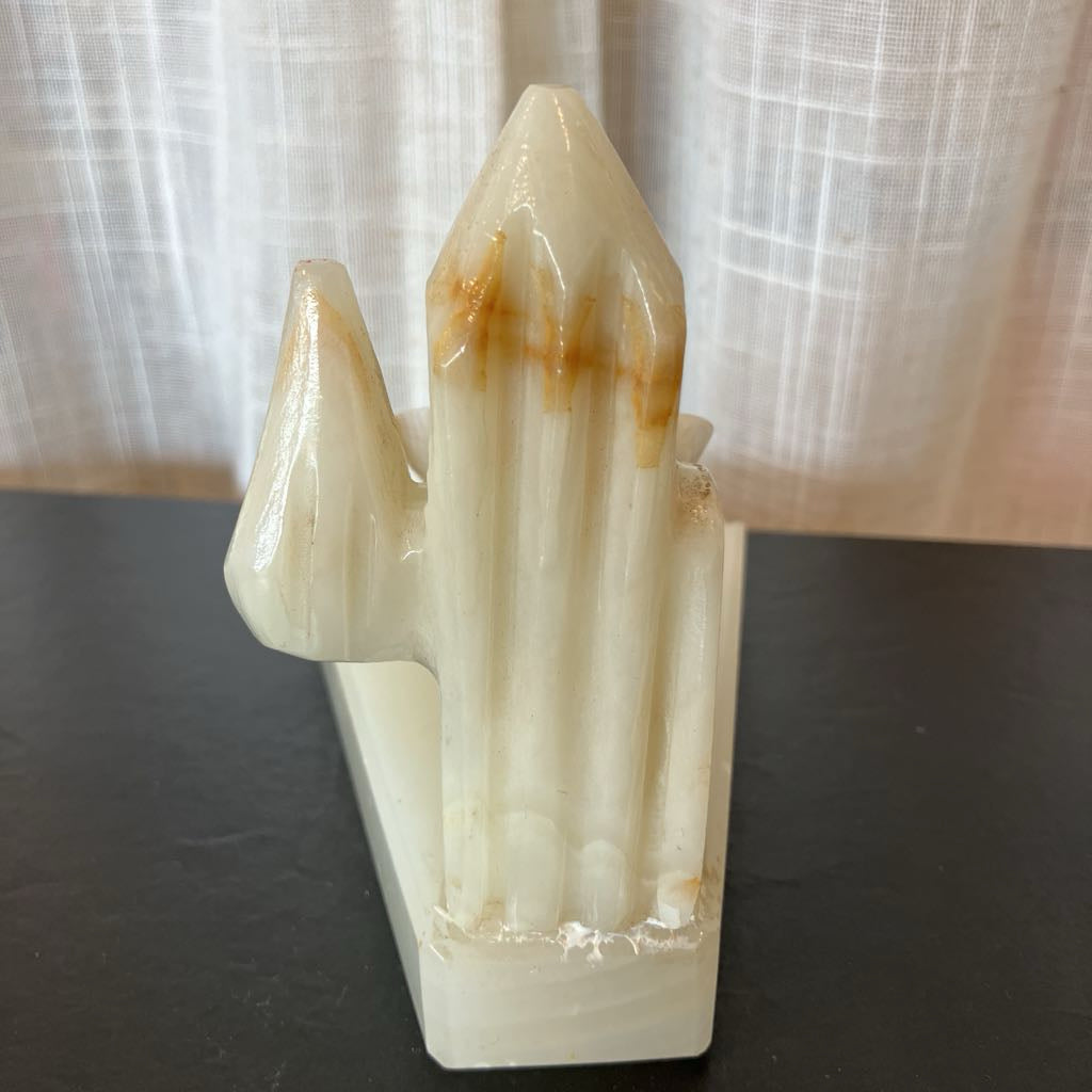 A carved stone crystal cluster figurine in off-white with natural brown and tan veining, standing on a dark surface against a light curtain backdrop.
