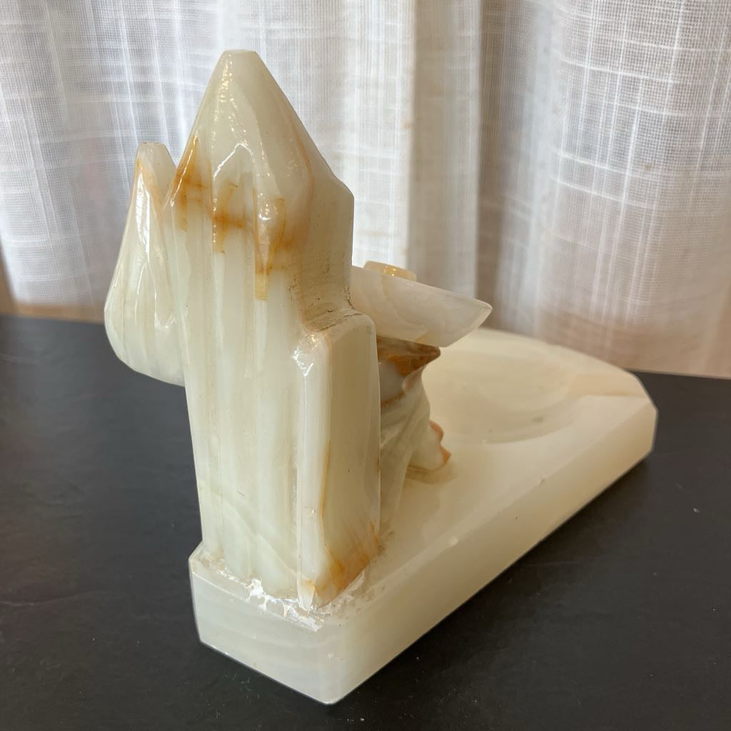A carved natural onyx stone sculpture depicting a stylized mountain or crystal formation on a rectangular base, photographed on a dark tabletop with sheer curtains in the background.