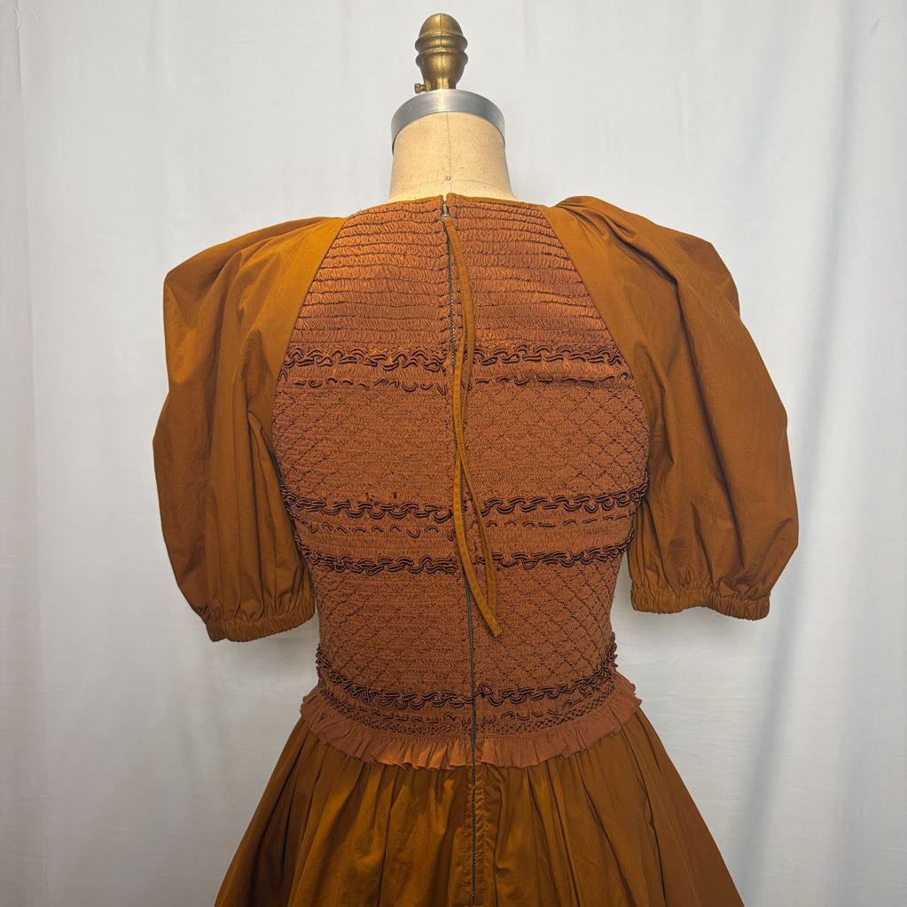 A vintage-style brown dress displayed on a dress form mannequin against a white curtain background. The back view shows a smocked bodice with horizontal gathered bands, a central tie, and voluminous puff sleeves.