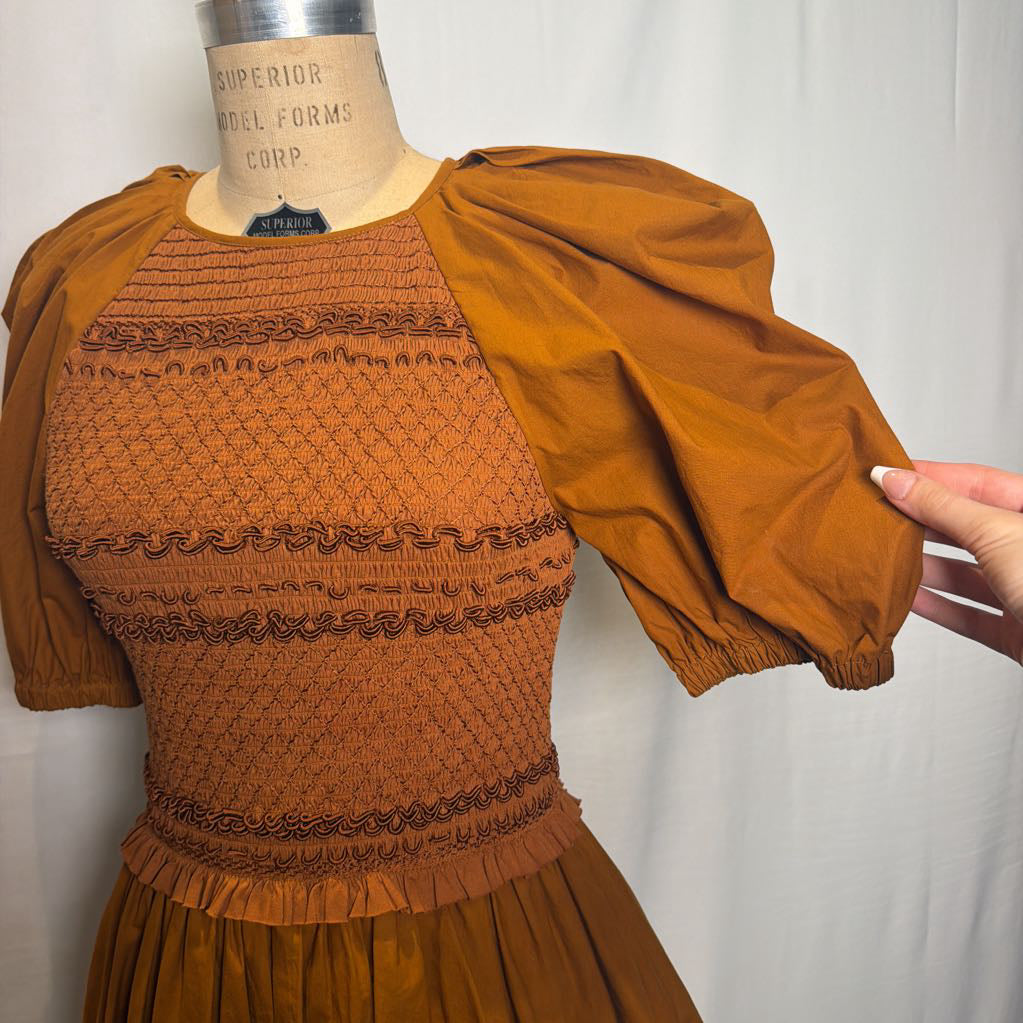 A vintage-style brown dress displayed on a mannequin, featuring a smocked bodice, puffed short sleeves, and a gathered skirt. A hand is holding out one of the sleeves to show its texture and shape against a plain white backdrop.