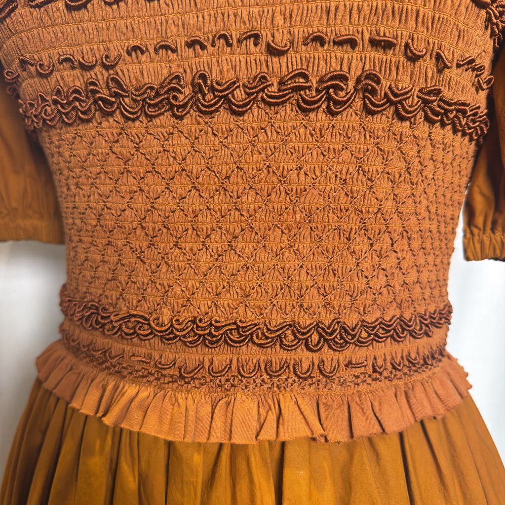 Close-up of a smocked dress bodice in a warm rust-orange color, featuring intricate textured stitching, ruffled trim, and a matching pleated skirt.