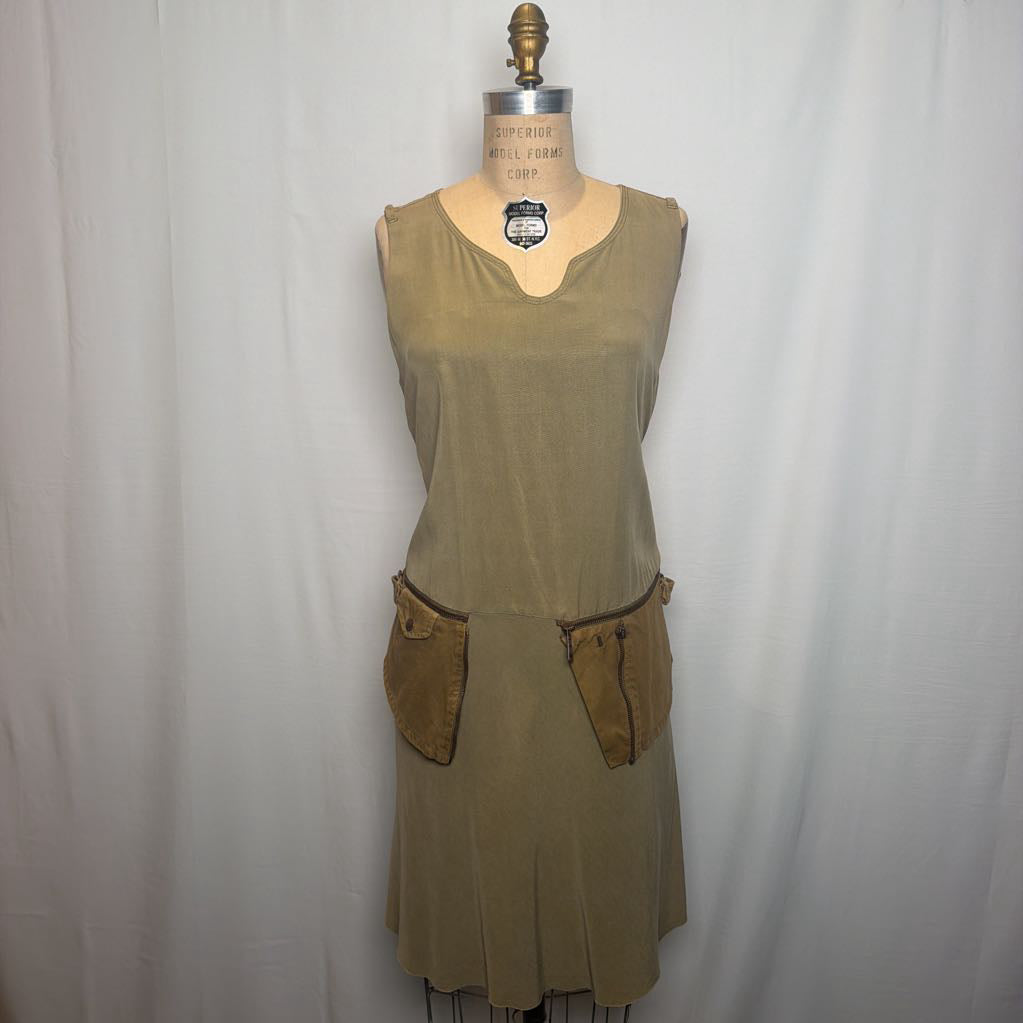 A sleeveless olive green dress displayed on a dress form mannequin against a white fabric backdrop. The dress features a split neckline and two large brown patch pockets on the front.