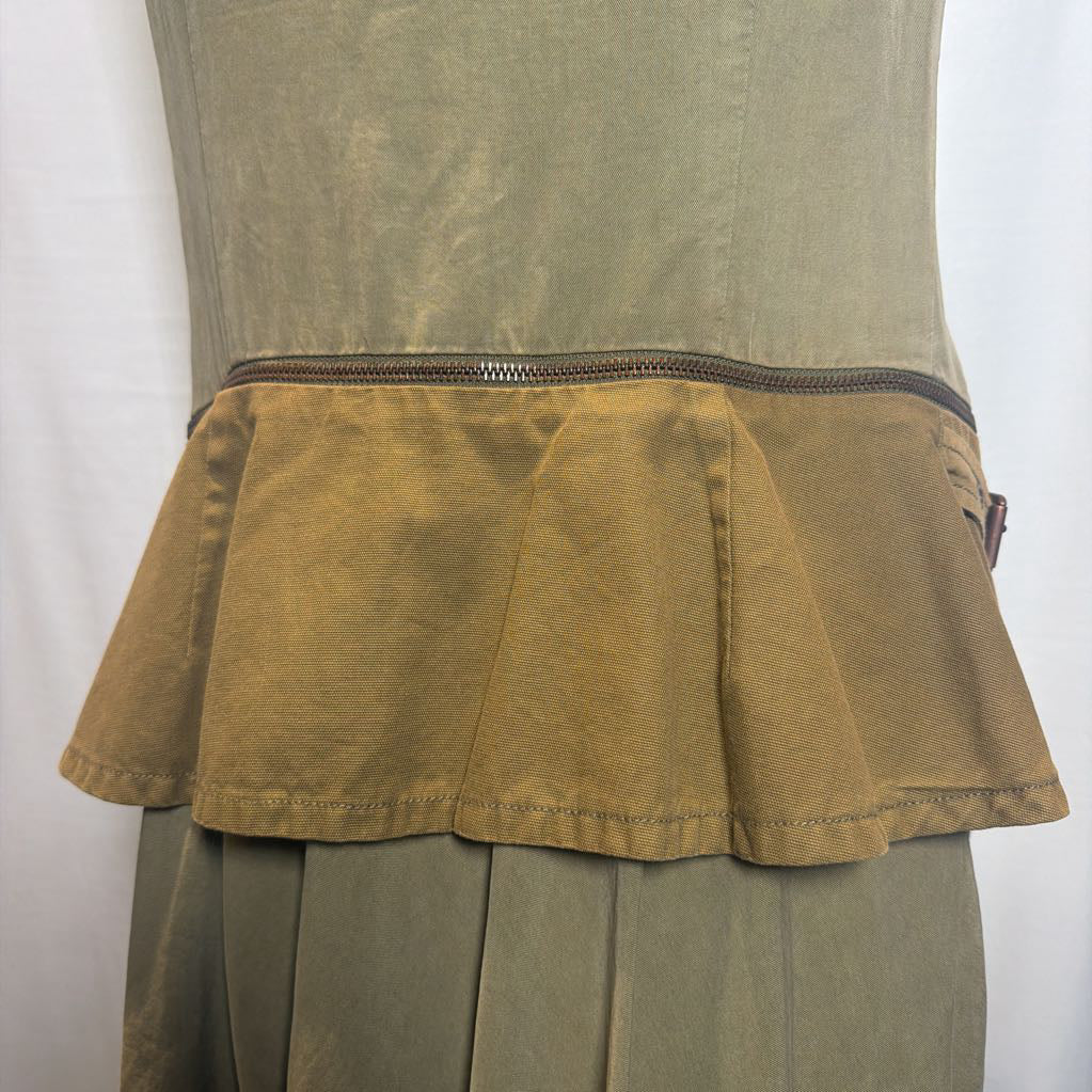 Close-up of a garment on a mannequin showing a pleated tan fabric waist detail with a dark zipper, attached to an olive green top and skirt.