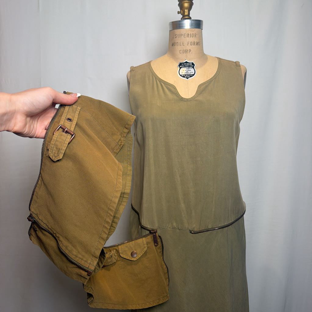 An olive green sleeveless dress displayed on a wooden mannequin, with a hand holding matching olive green shorts and a matching belt pouch against a light gray fabric backdrop.
