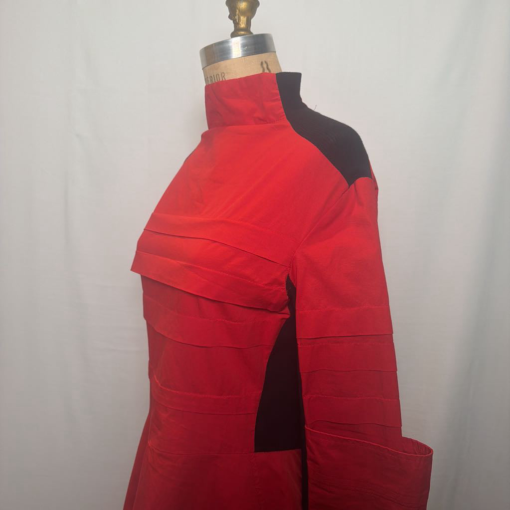 A red and black high-neck long-sleeve garment displayed on a dress form mannequin against a white fabric backdrop.
