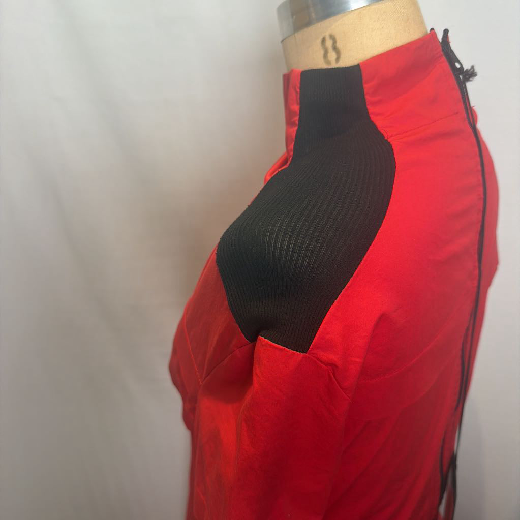 A close-up side view of a red athletic jacket displayed on a dress form. The jacket features a black ribbed knit shoulder yoke and a black side zipper.
