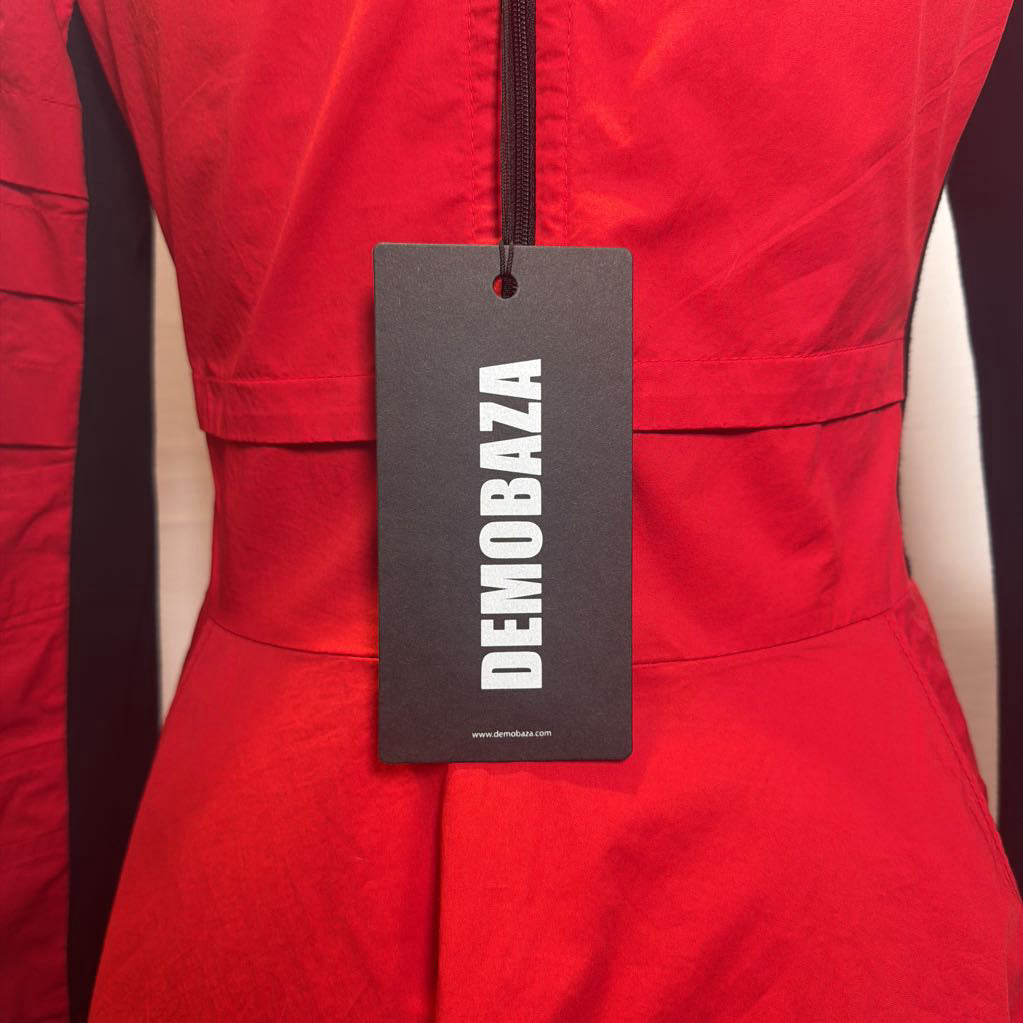 A close-up of a bright red garment with a black hangtag hanging from the zipper. The tag is printed with the brand name 'DEMOBAZA' in white capital letters.