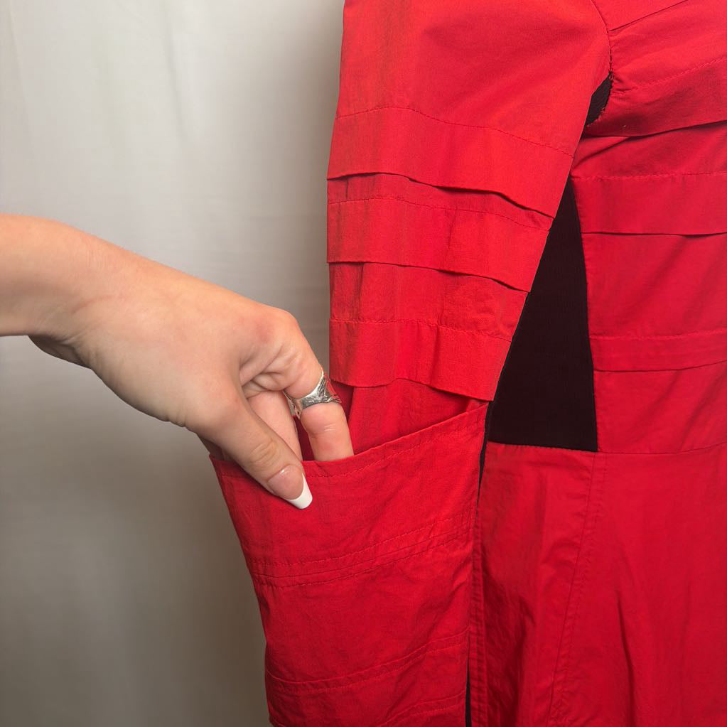 A close-up of a person's hand with a French manicure reaching into a side pocket of a bright red work jacket or bib, showing a silver ring on the finger.
