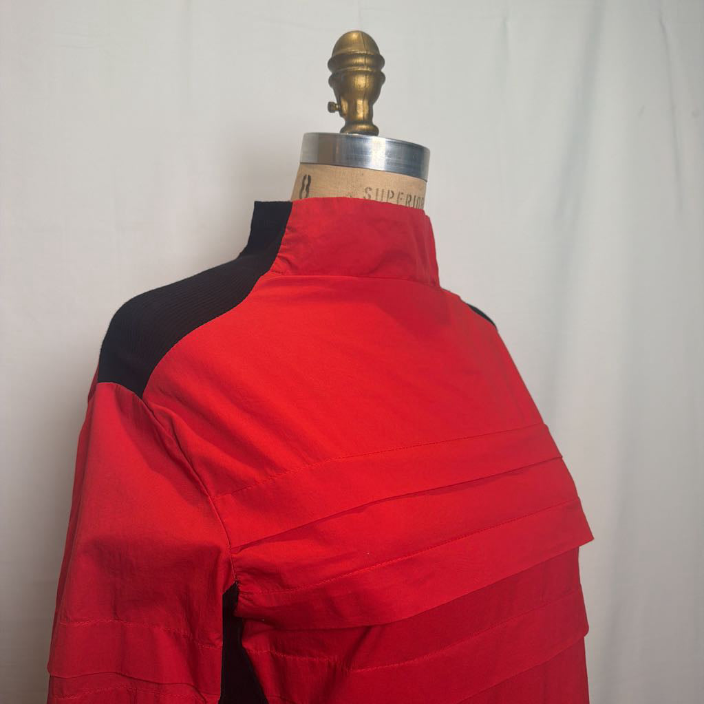 A red and black long-sleeve top displayed on a dress form mannequin against a white fabric backdrop.