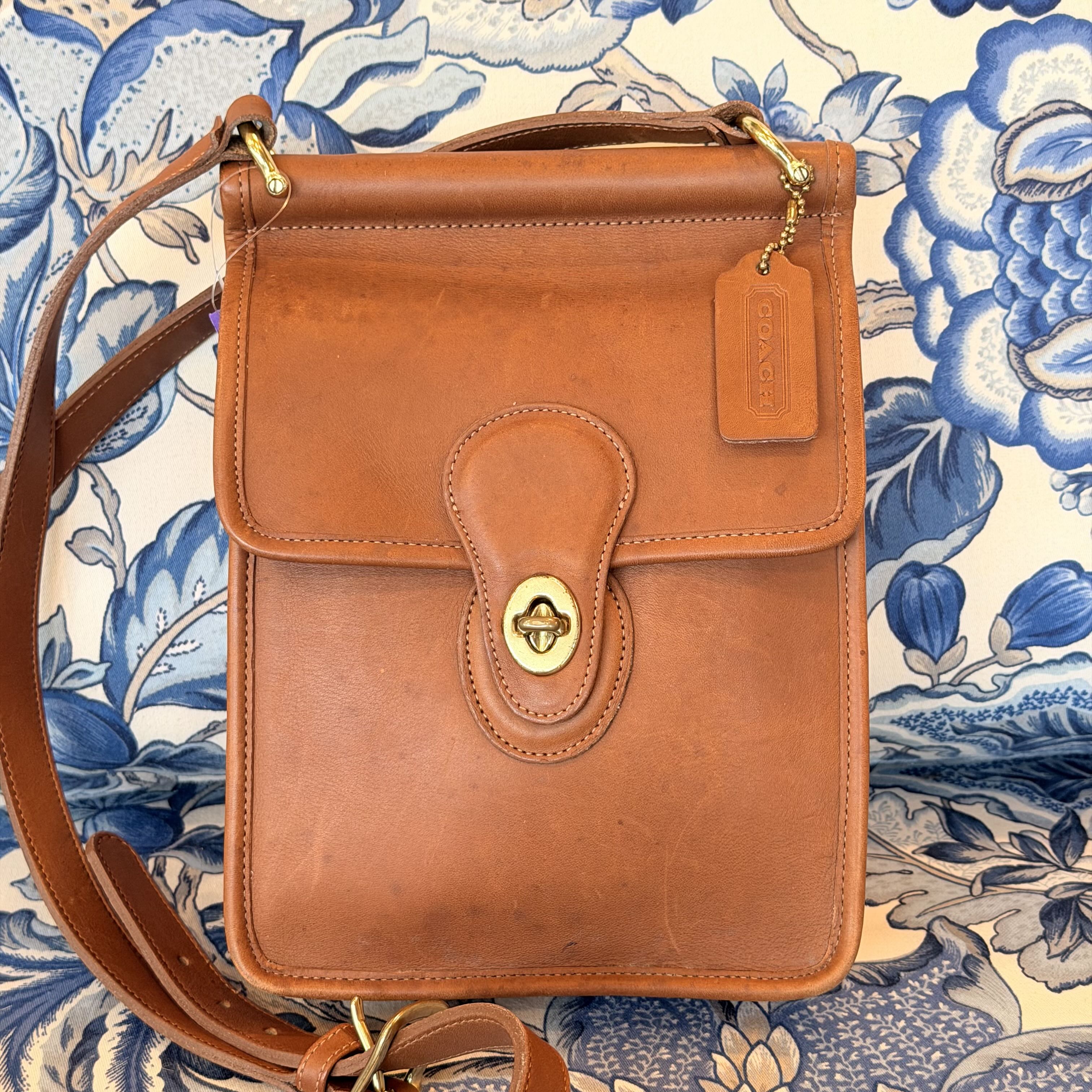 A vintage brown leather crossbody bag with a gold-tone turn-lock clasp and a hanging leather tag, laid flat on a blue and white floral patterned fabric background.