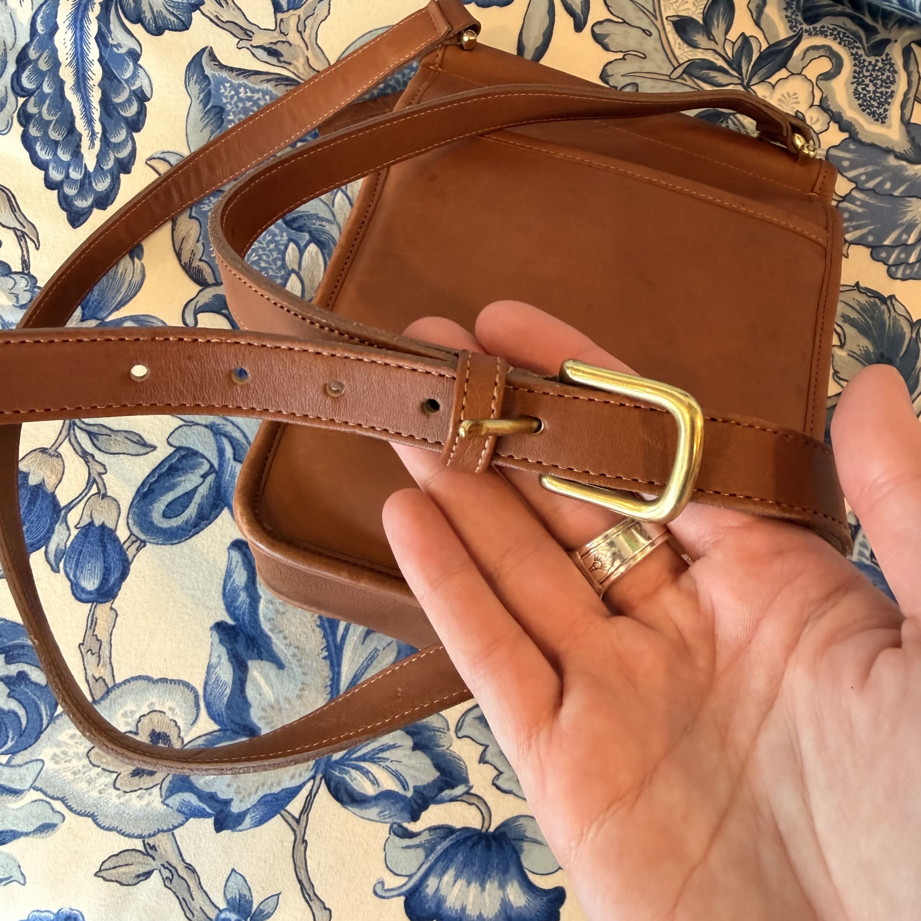 A brown leather shoulder bag with a matching belt strap and gold-tone buckle, held by a hand over a blue and cream floral patterned fabric background.