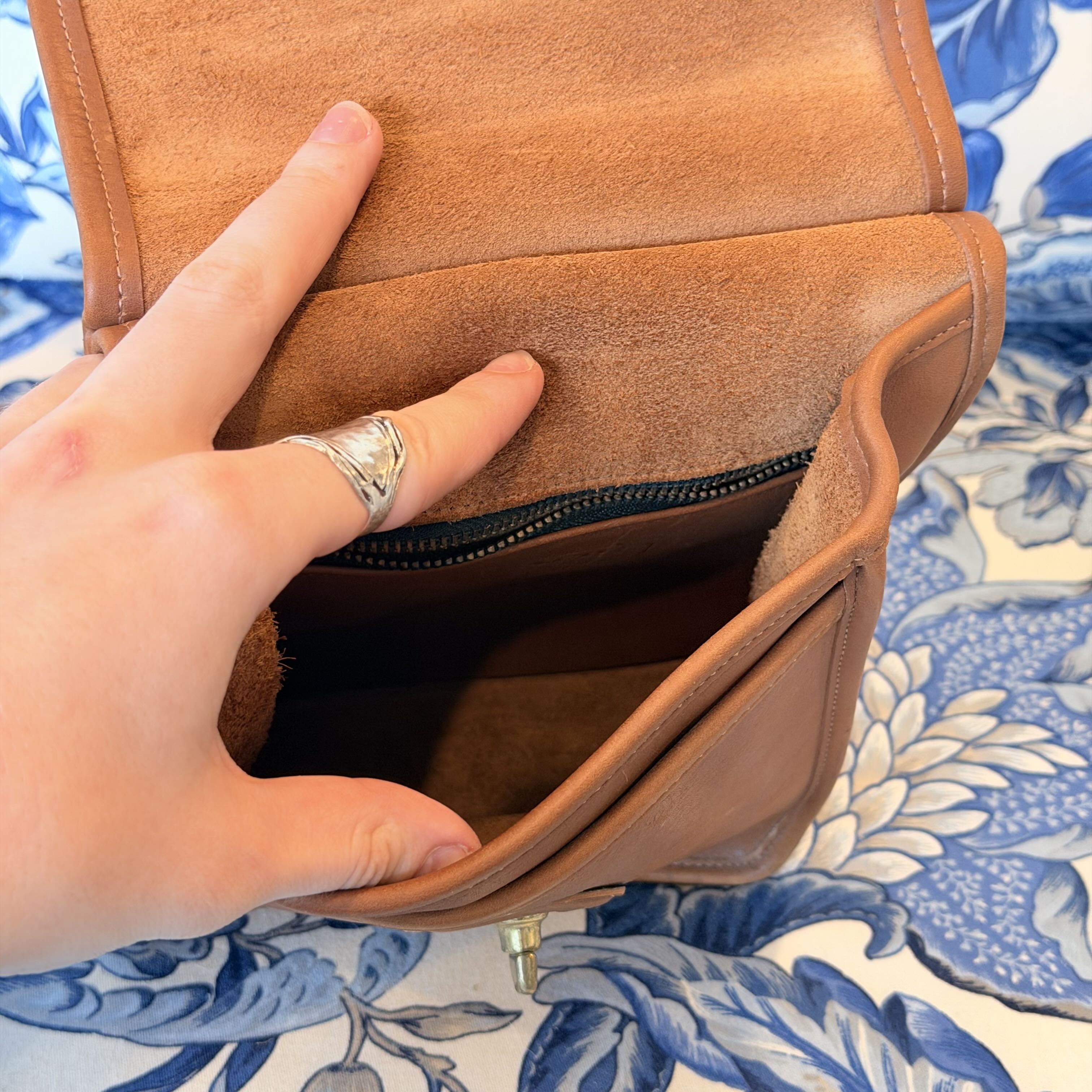 A close-up of a person's hand opening a tan suede or faux-suede clutch or wallet, showing the interior zip pocket, against a blue and white floral fabric background.