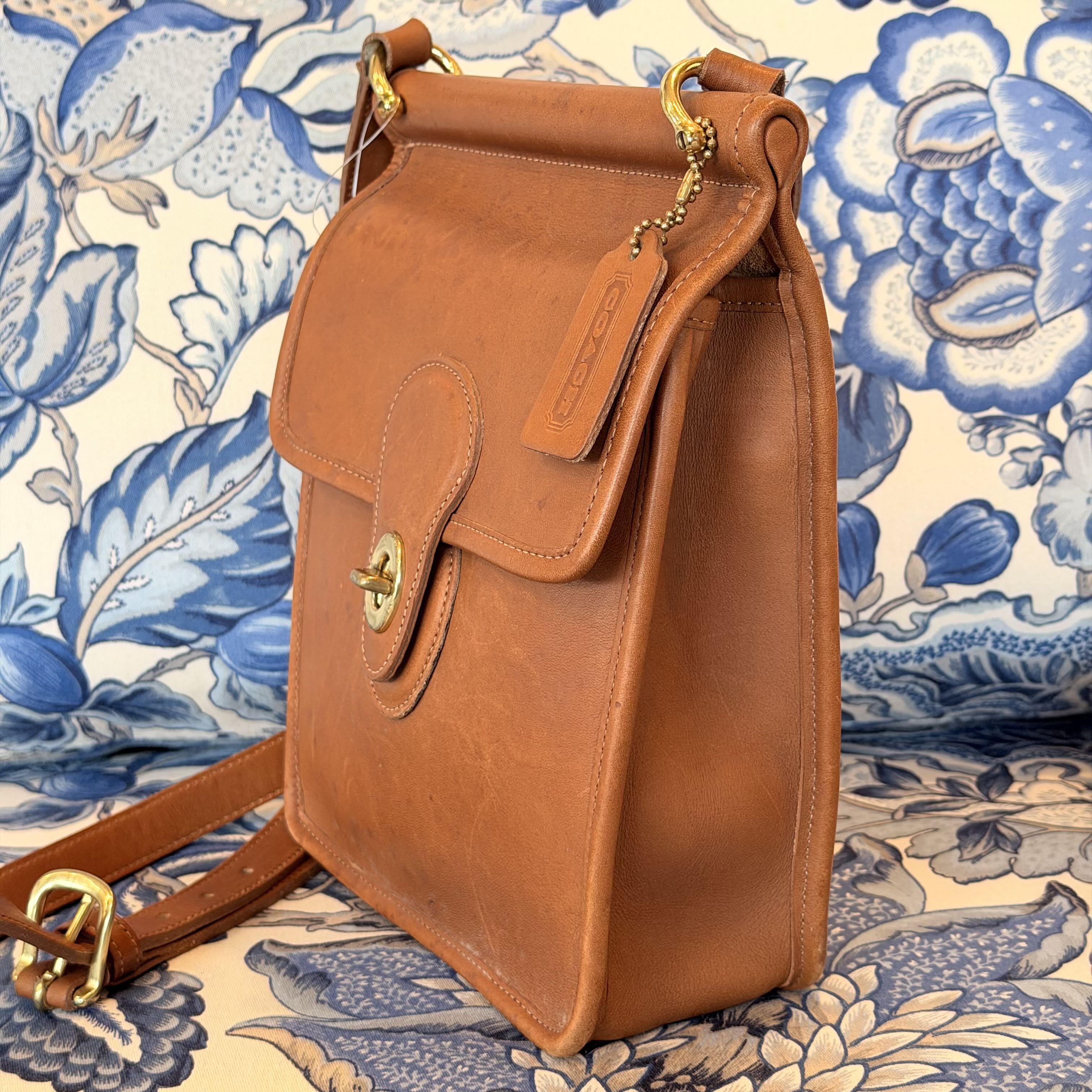 A tan leather shoulder bag with a front flap and gold-tone hardware, photographed against a blue and cream floral patterned fabric background.