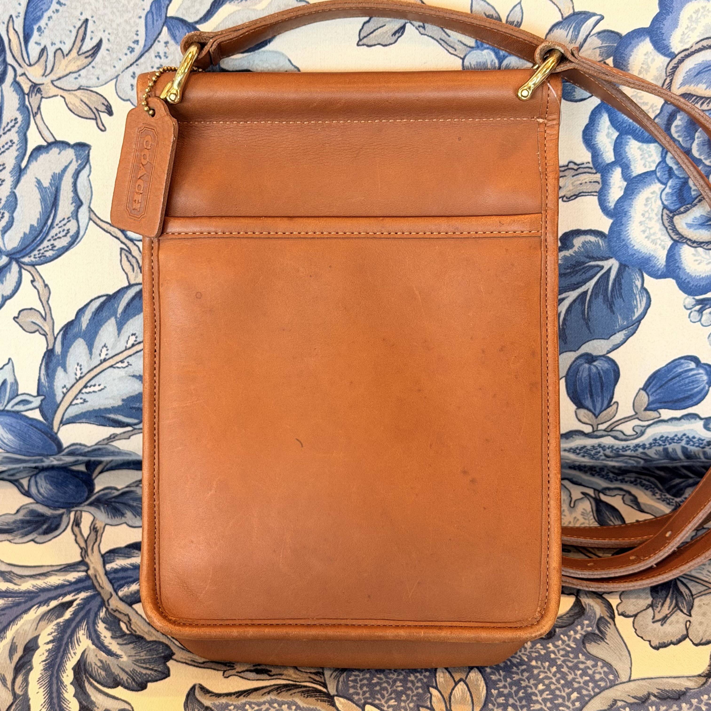 A tan leather crossbody bag with gold-tone hardware, laid flat on a blue and cream floral patterned fabric background. The brand name 'DIXIE' is embossed on the leather hangtag.