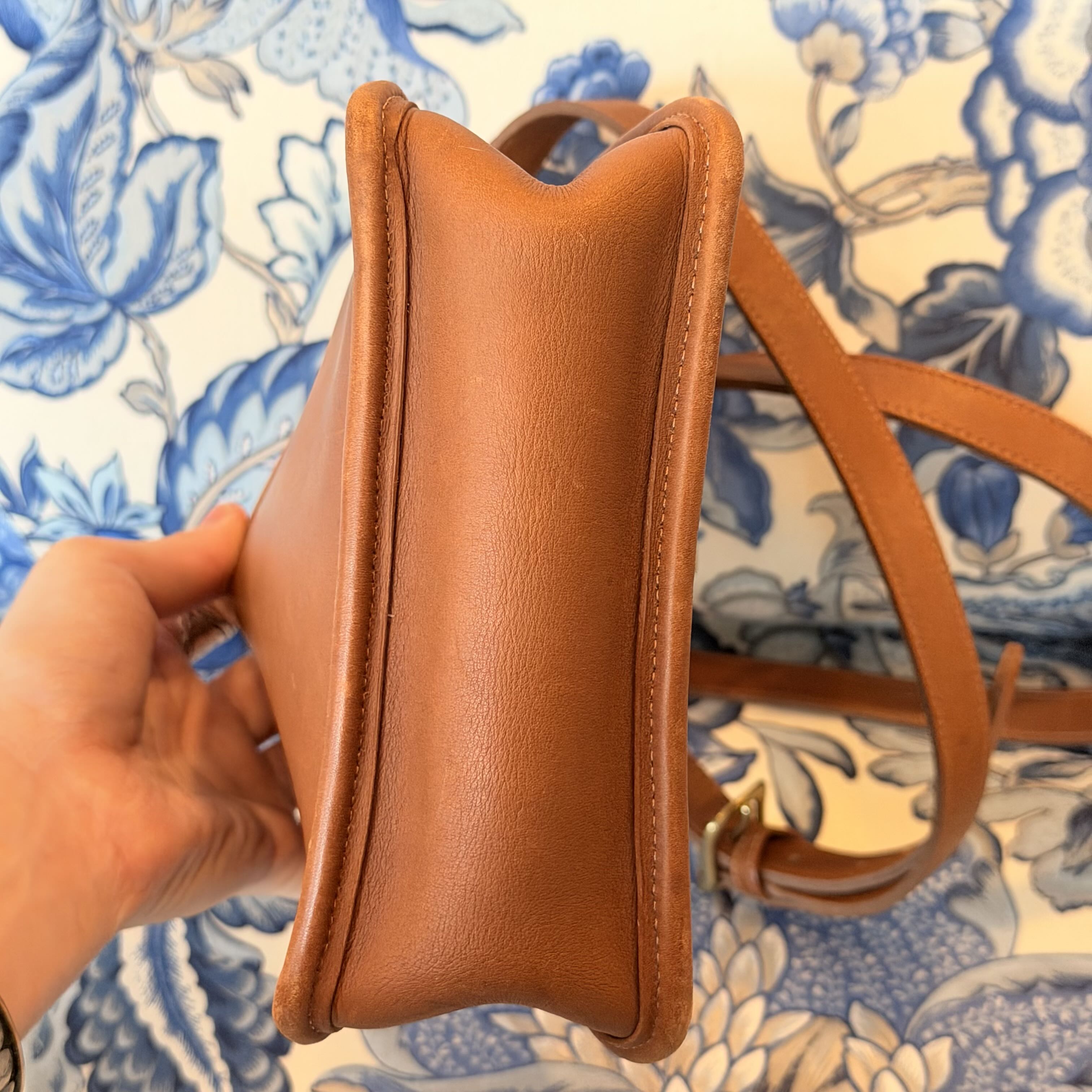 A close-up of a tan leather shoulder bag being held by a hand, showing the side profile and stitching, with a blue and white floral patterned fabric in the background.