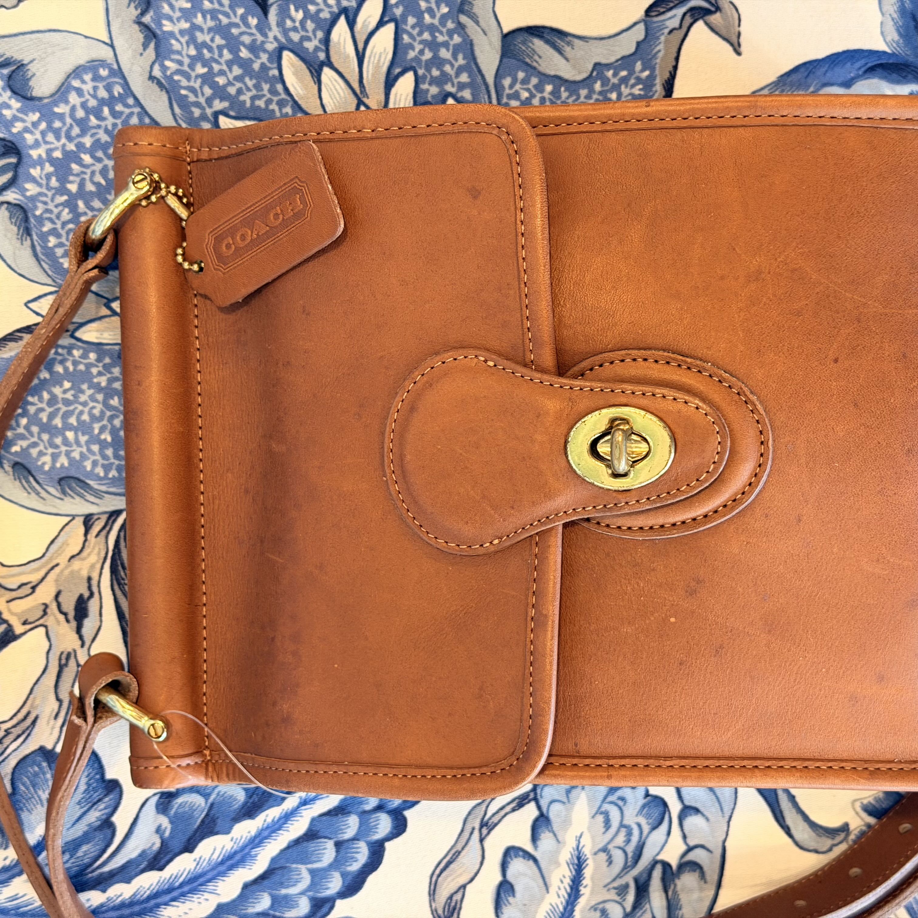 A Coach leather book cover or document holder in a warm cognac brown, featuring a snap-button closure, a leather brand tab, and gold-tone hardware, laid flat on a blue and cream patterned textile background.