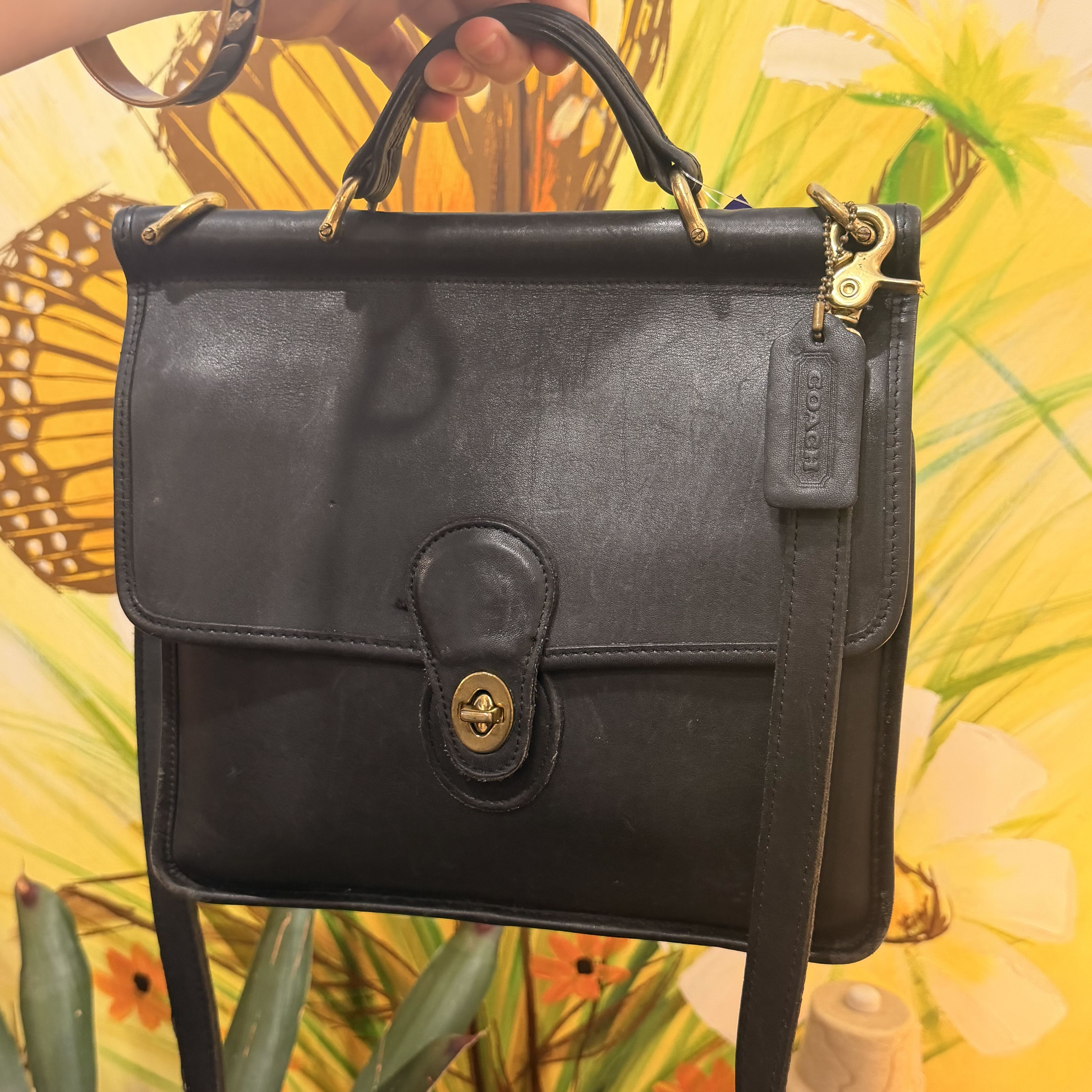 A black leather Coach satchel bag with gold-tone hardware, held by a hand against a vibrant yellow tropical floral background.