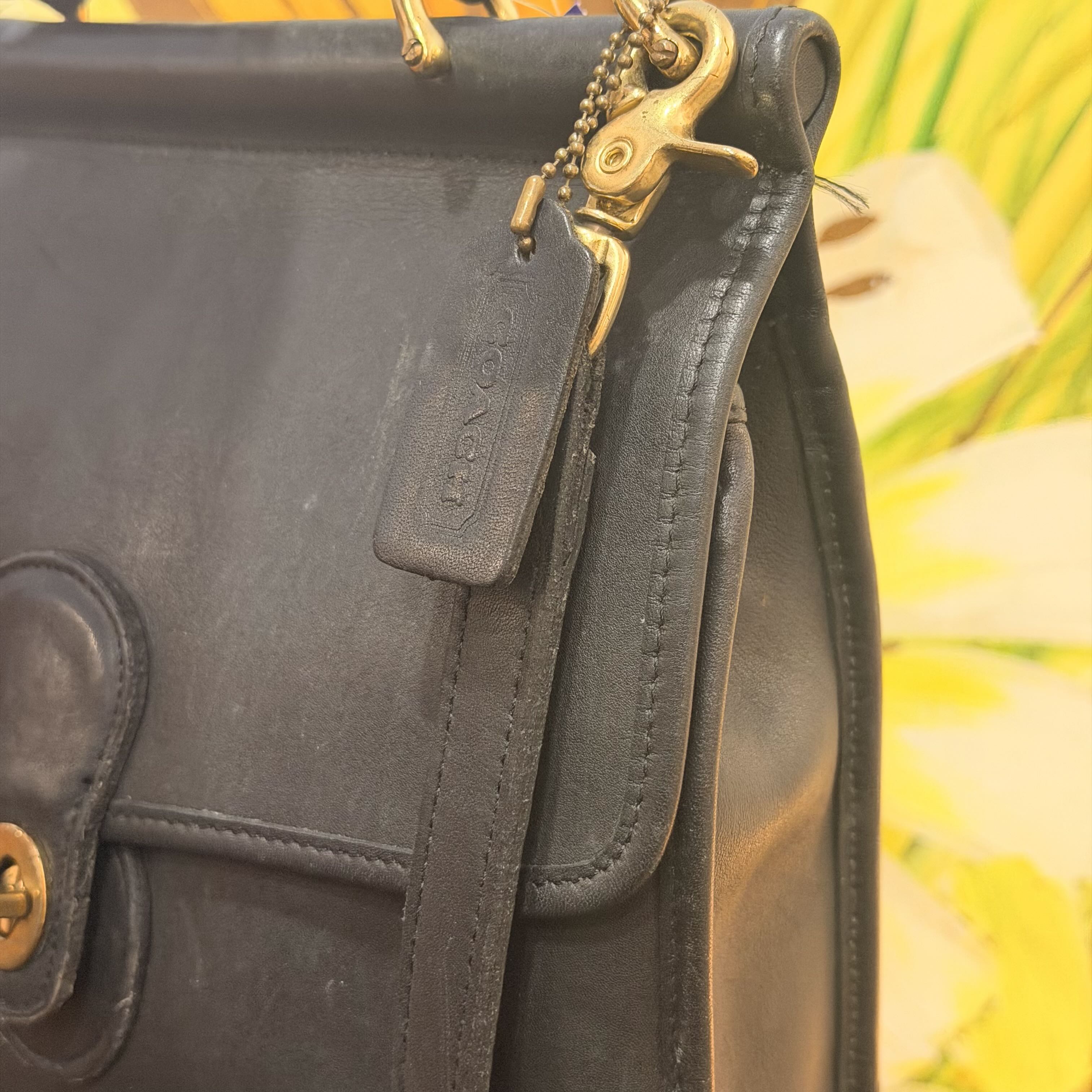 Close-up of a black leather Coach handbag with gold-tone hardware, a hanging leather tag embossed with Coach, and a blurred yellow-green floral background.