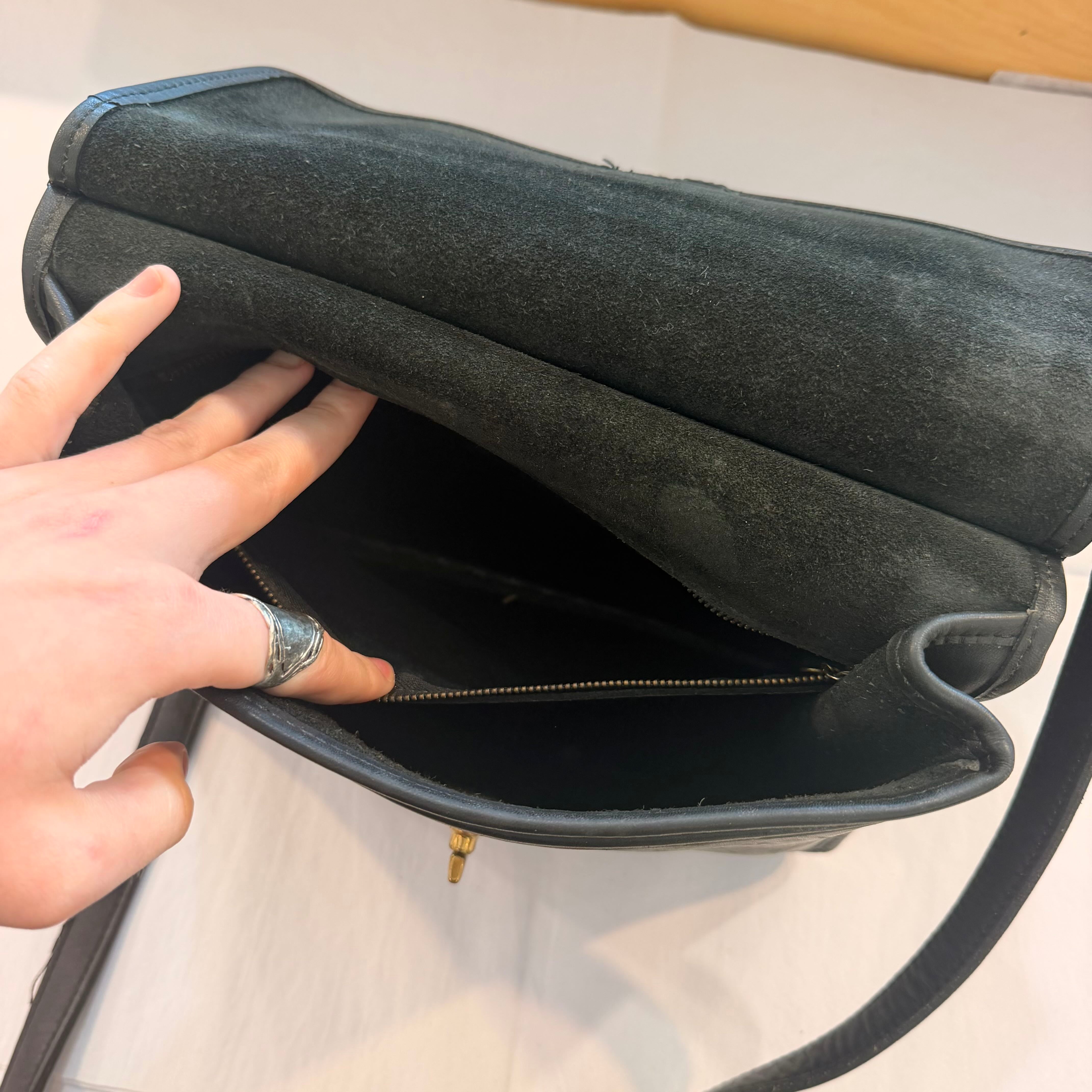 A close-up of a black suede shoulder bag being opened by a hand, showing the interior zippered compartment and black leather trim.