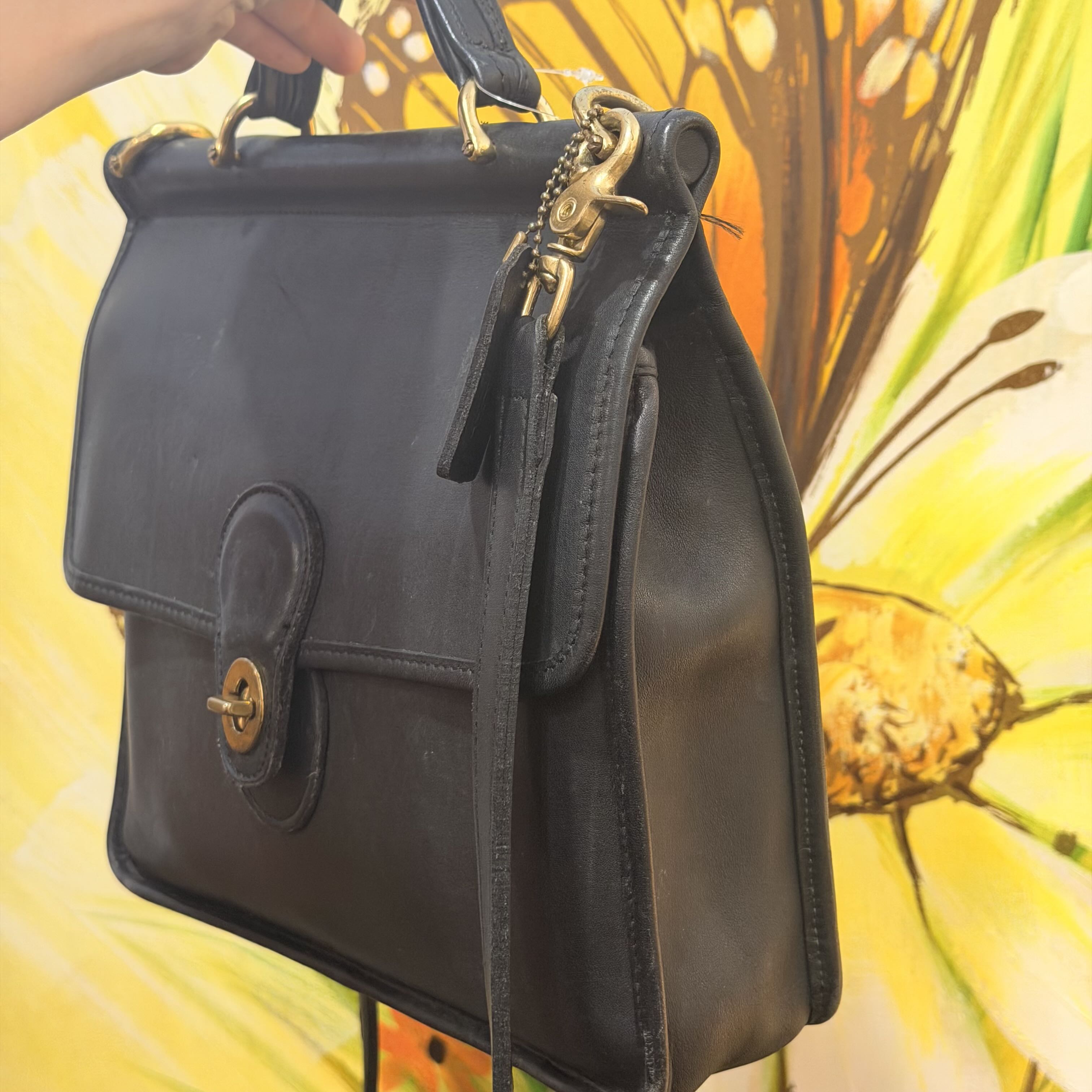 A black leather handbag with gold-tone hardware, held by a hand against a bright yellow and orange floral background.