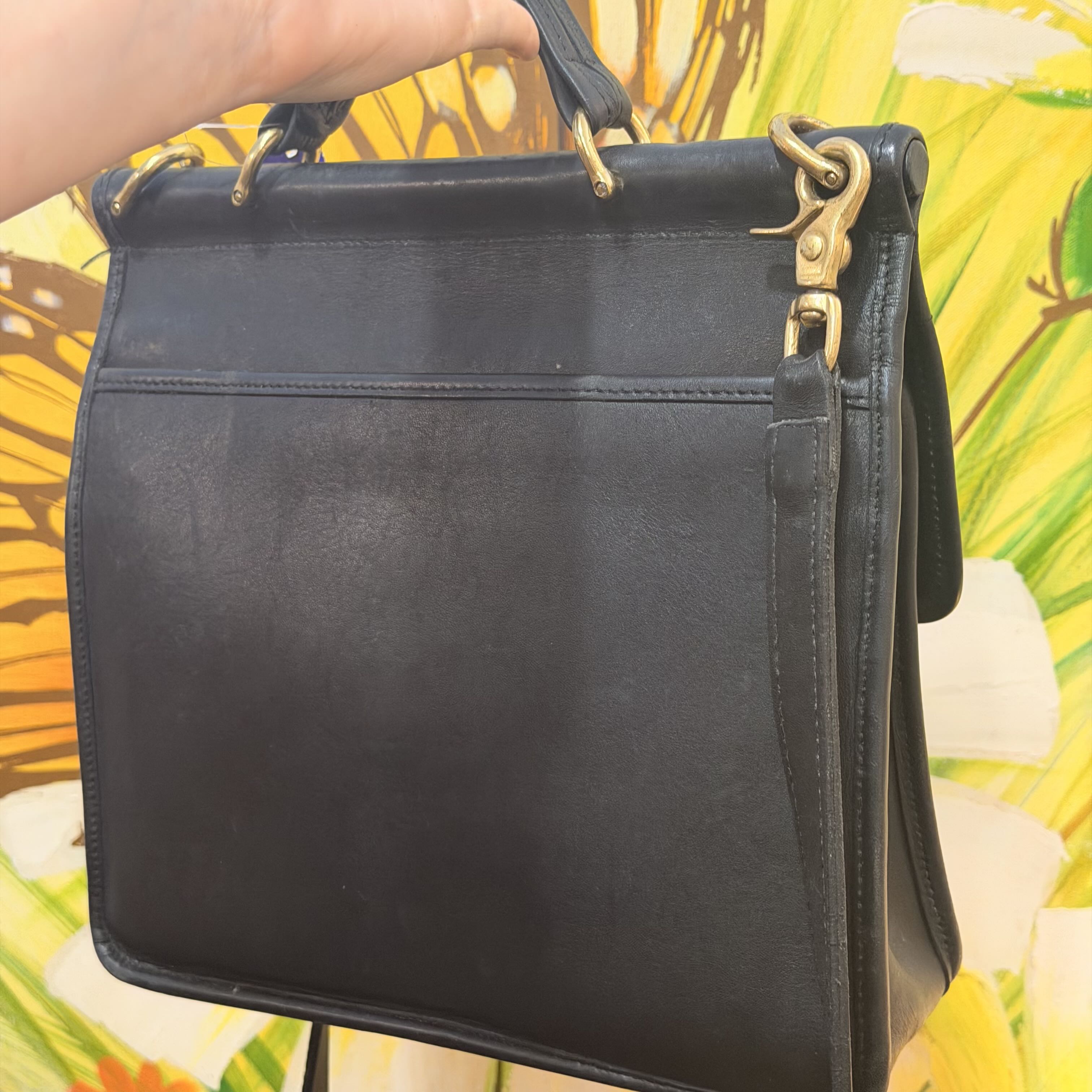 A black leather handbag with gold-tone hardware, held by a hand against a bright yellow tropical floral background.