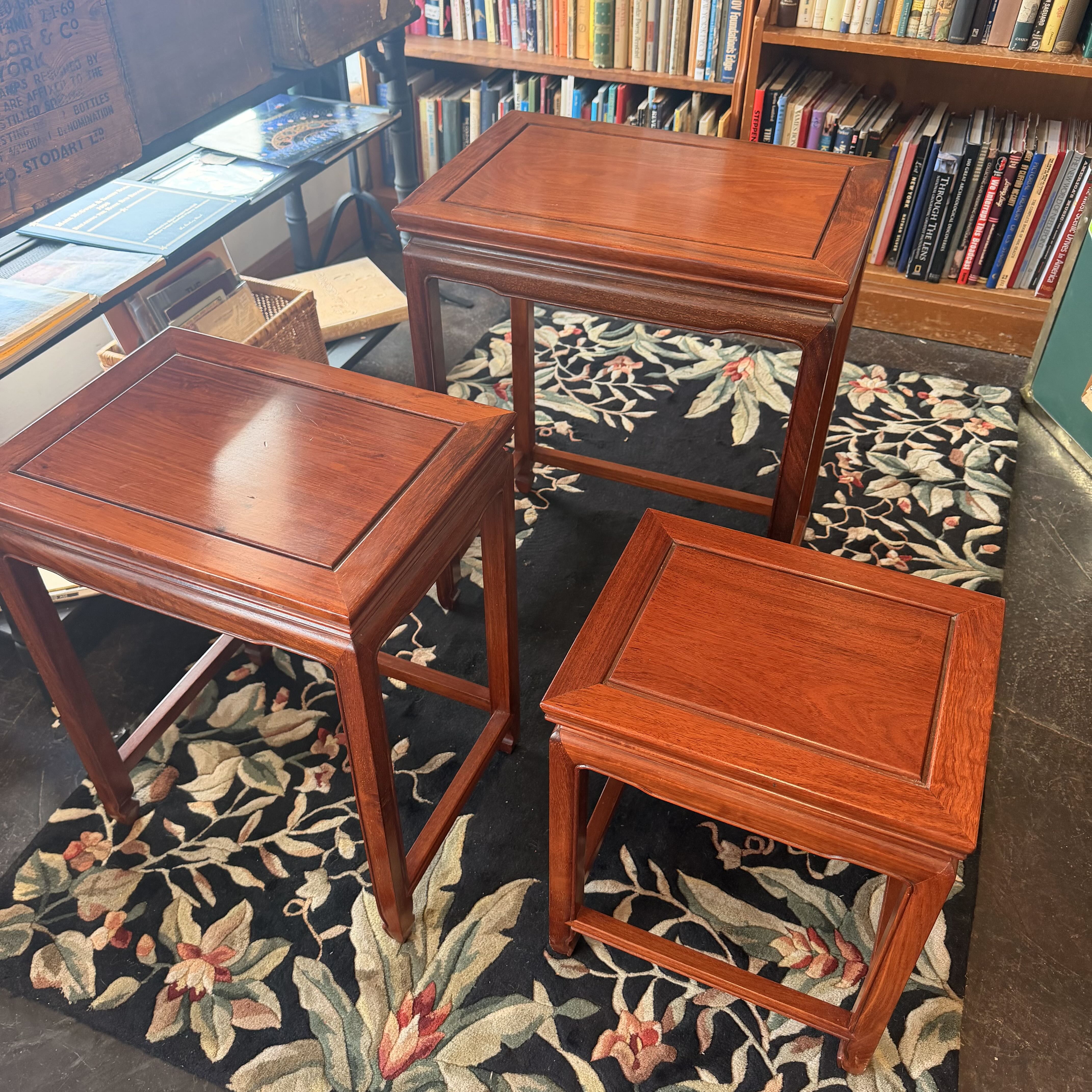 Three reddish-brown wooden nesting tables or small side tables arranged on a dark floral rug in a book-filled room.