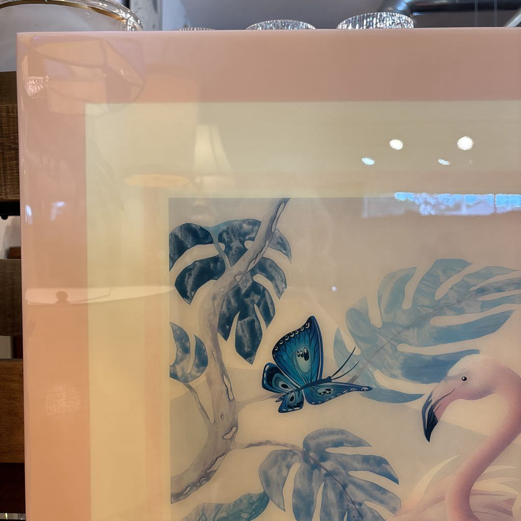 A framed tropical art print featuring blue monstera leaves, a blue butterfly, and a pink flamingo, displayed in a glossy frame with warm reflections.